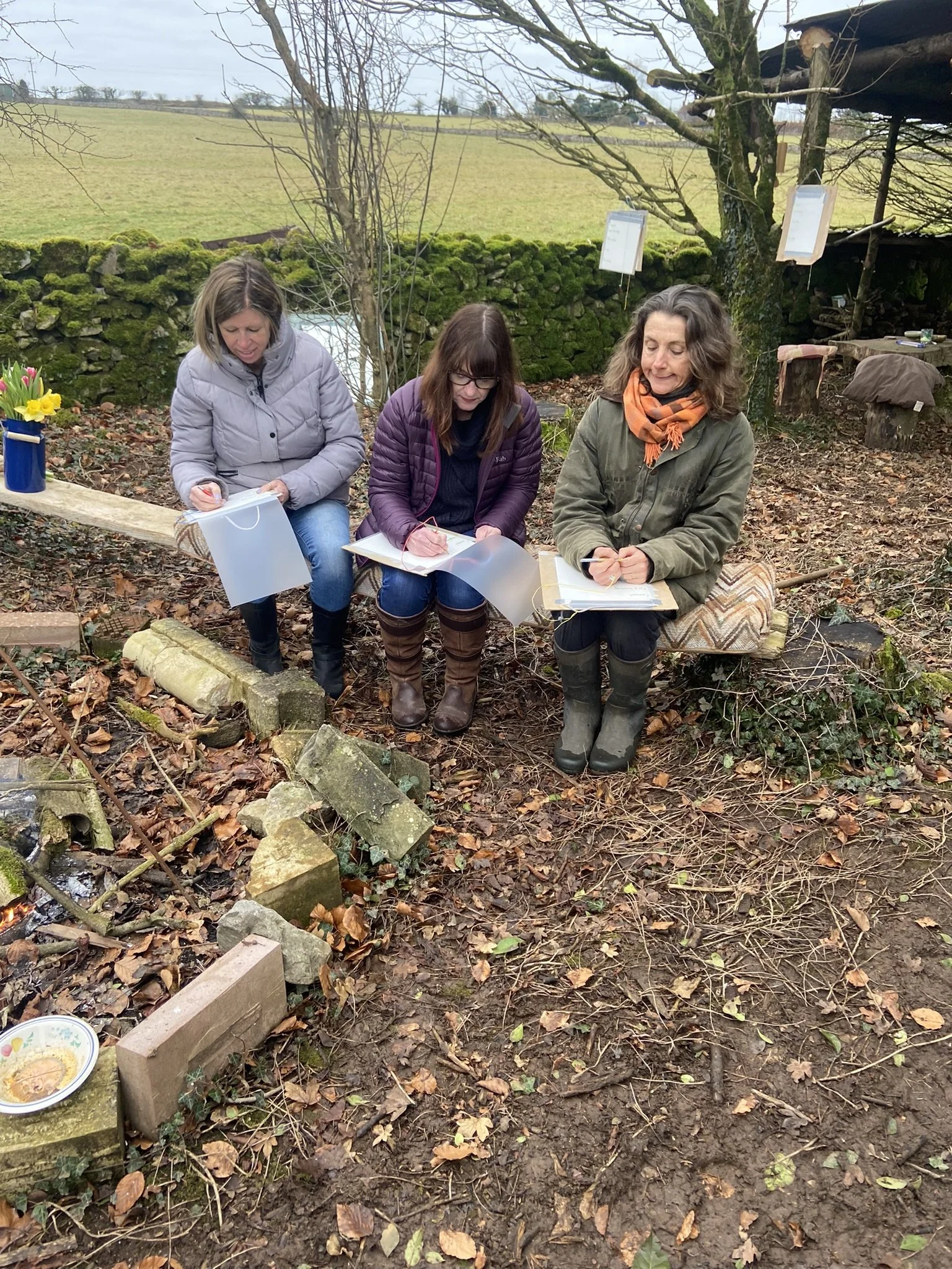 Three women sitting outdoors on a wooden bench or platform, writing or taking notes on clipboards or papers, with a rural landscape in the background, moss-covered stone wall, tree, and various outdoor objects around them.