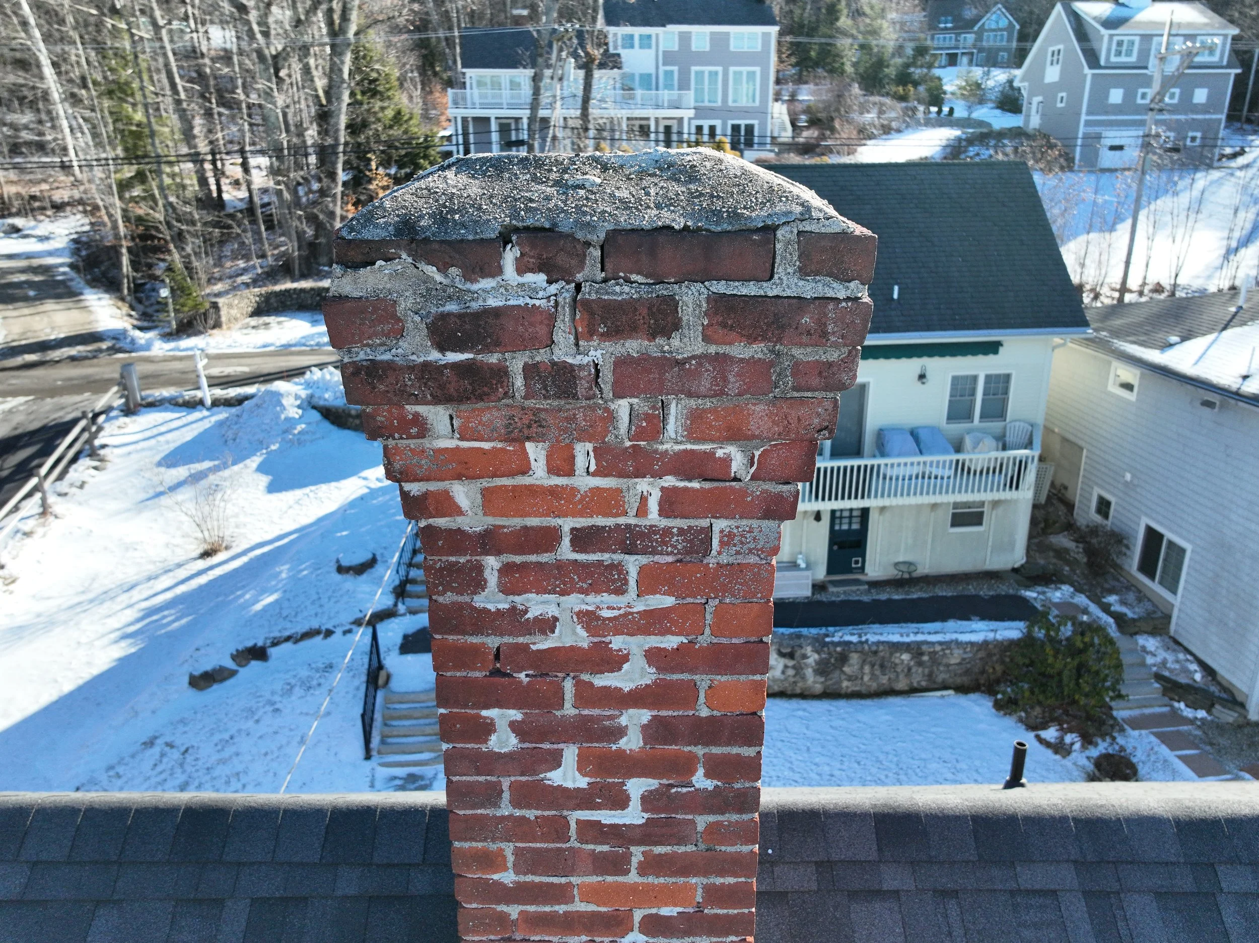 A brick chimney on a rooftop overlooking a snowy residential neighborhood with various houses and trees.