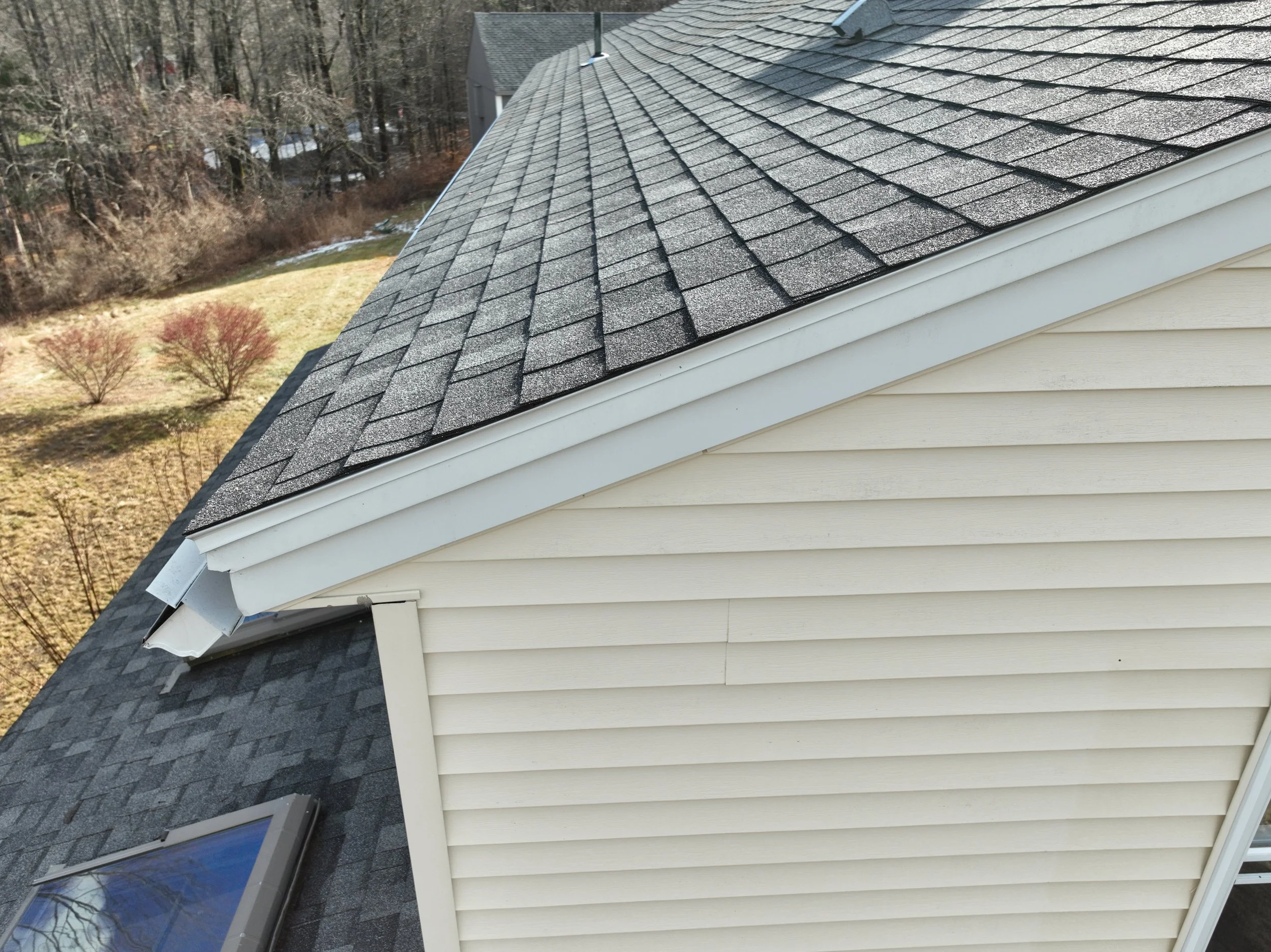 A side view of a house roof with gray asphalt shingles, white siding, and a skylight window, with trees and a lawn in the background.