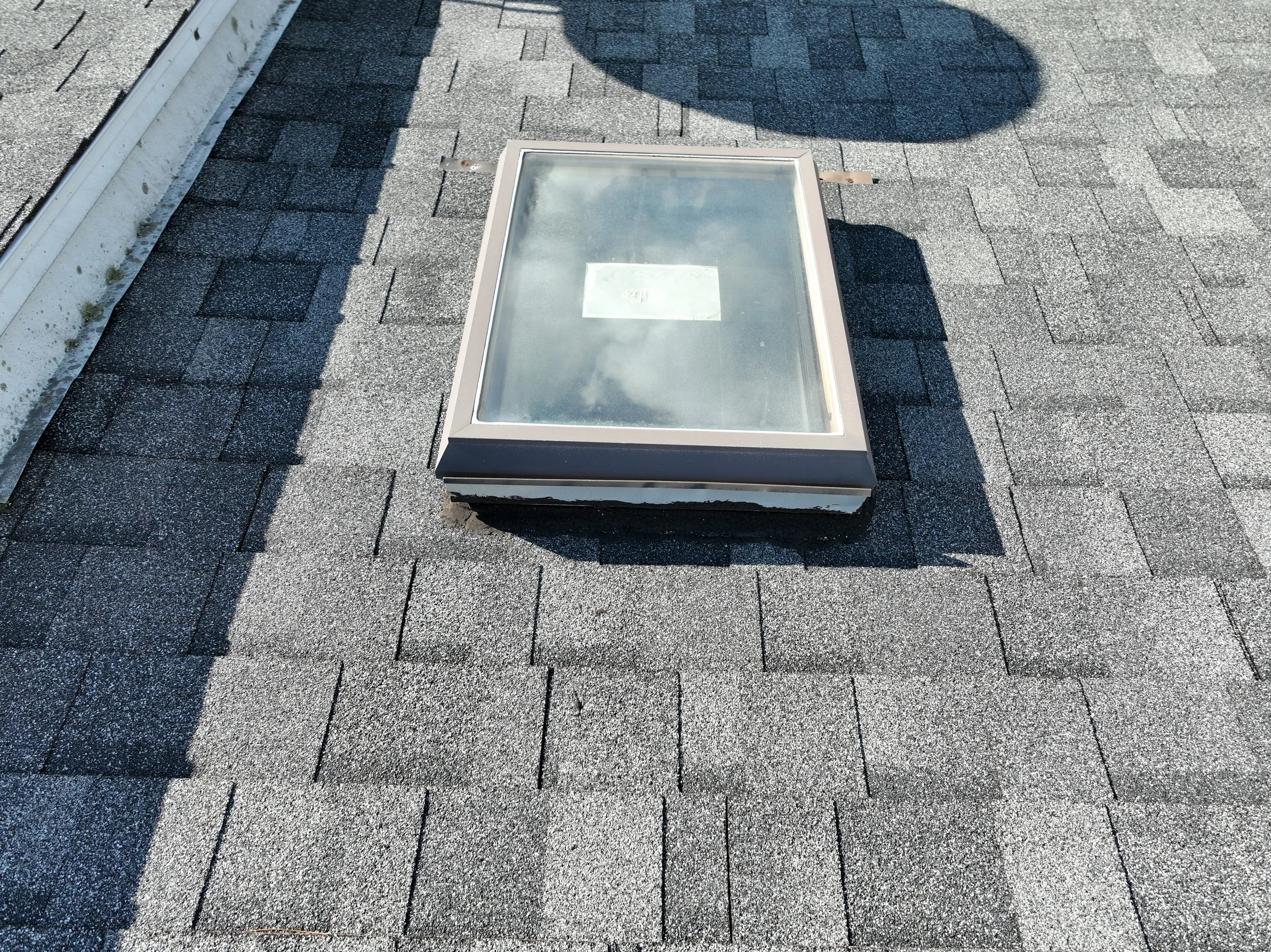 View of a skylight vent on a flat roof covered with asphalt shingles, with shadows cast by nearby structures.