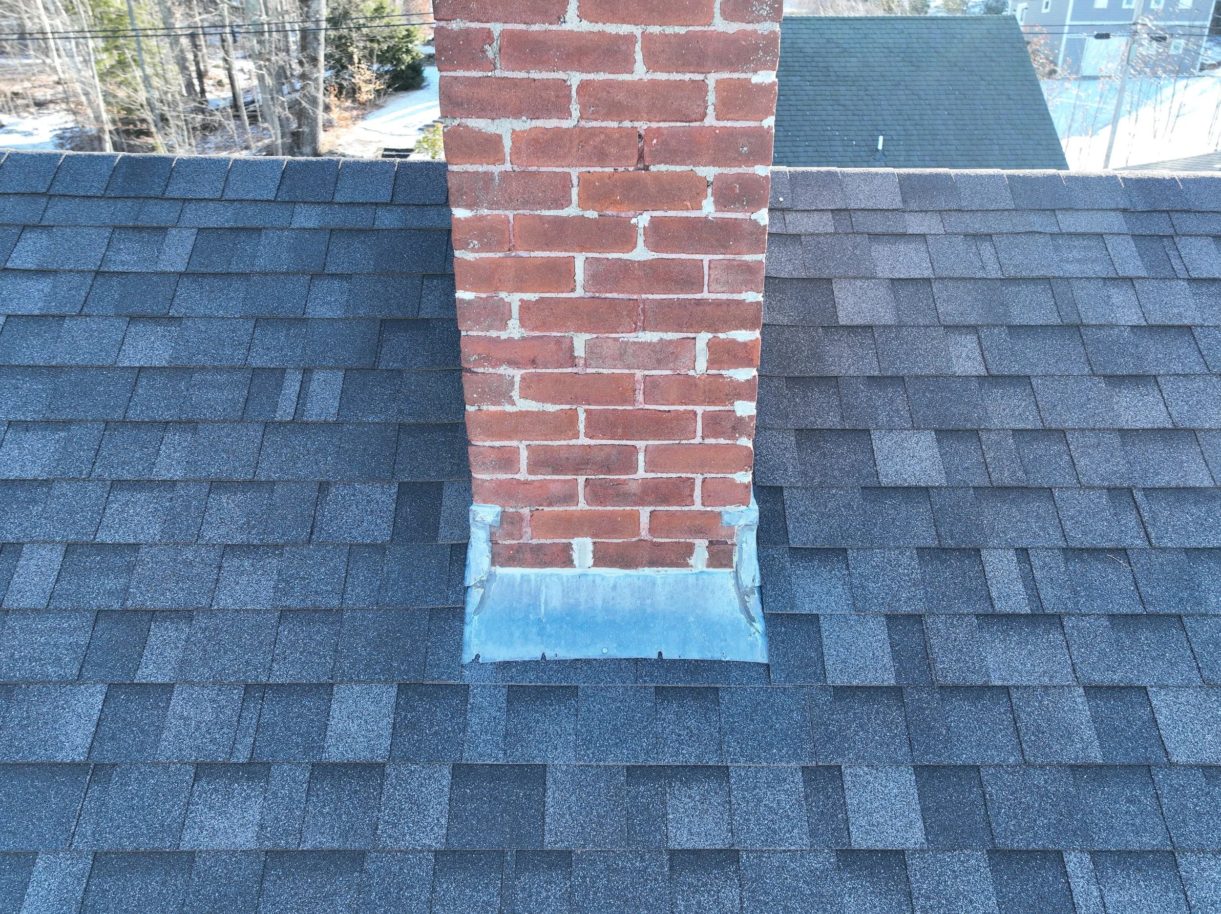 A rooftop view showing dark asphalt shingles and a brick chimney covered by metal flashing at the base, with trees and neighboring roofs in the background.