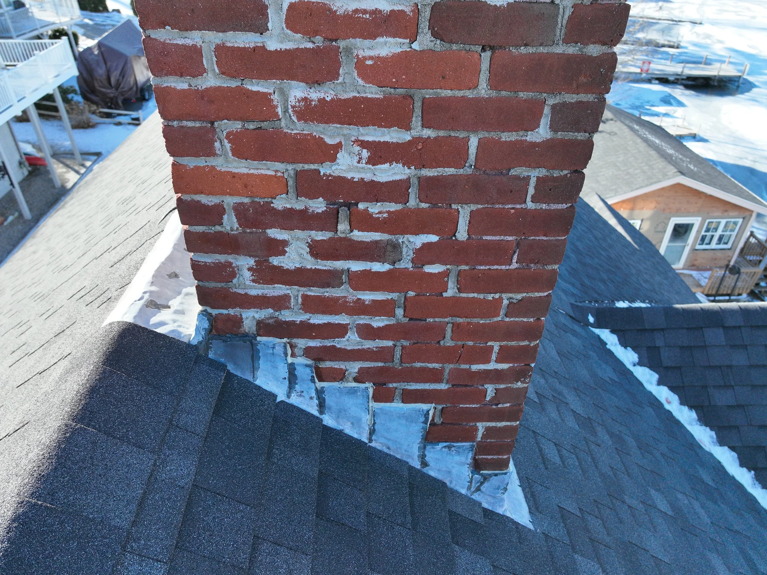 Close-up of a brick chimney on a house roof, with surrounding snow and other rooftops visible in the background.