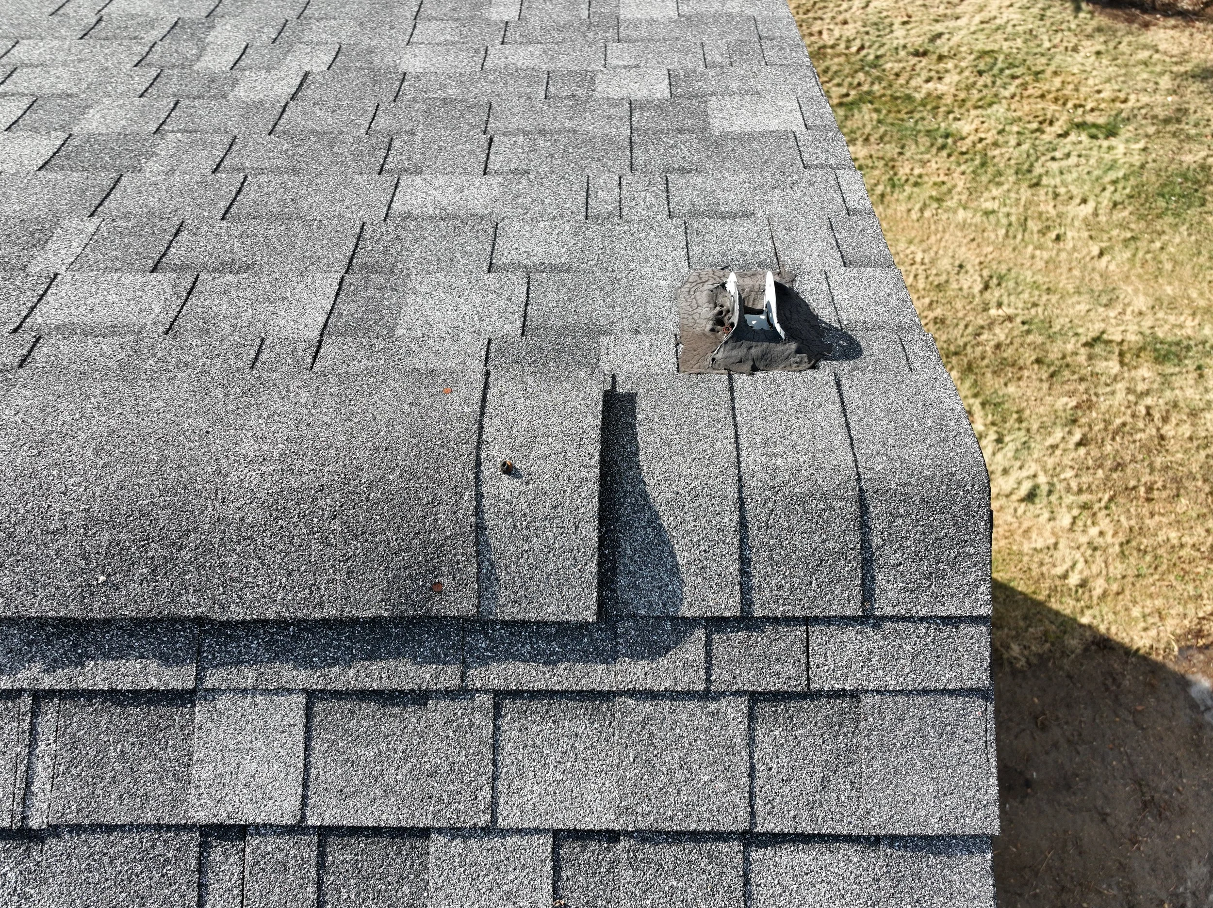 Close-up of a shingled roof with a hole and damaged shingles, and a shadow cast on the roof.