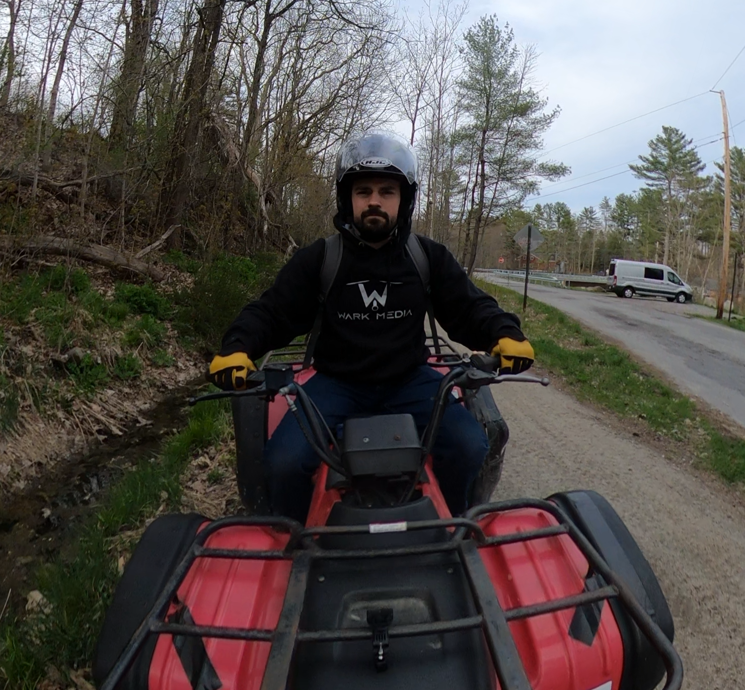 Man wearing a black hoodie with 'WARK MEDIA' logo, yellow gloves, helmet, riding a red ATV on a gravel road near trees and a parked white van.