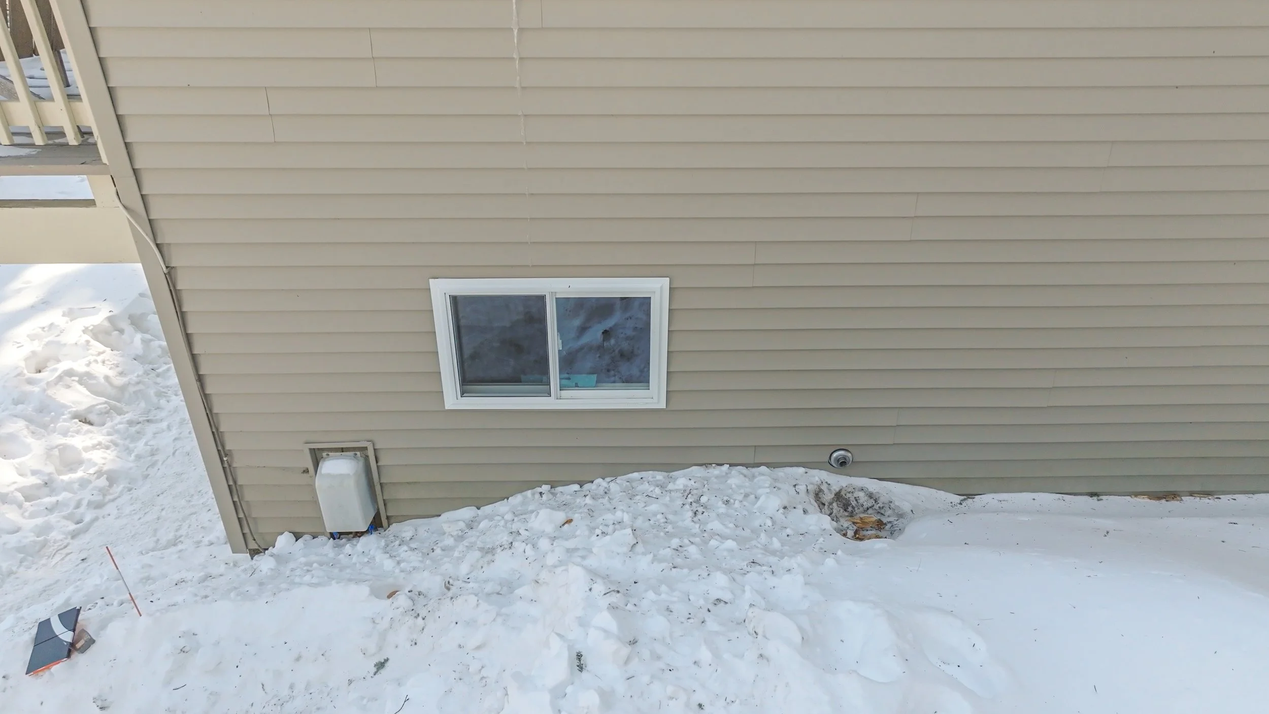 Side view of a beige house with a snow-covered ground, a small window, an outdoor electrical box, and a vent.