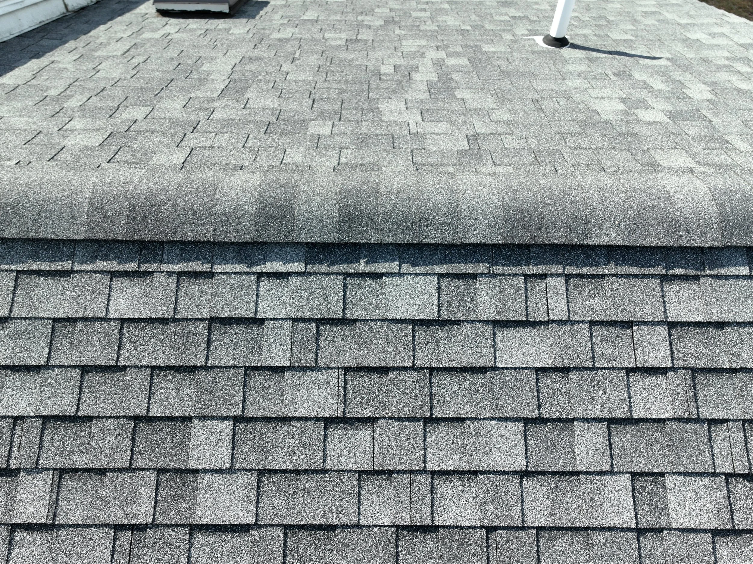 A rooftop showing different sections of asphalt shingles, with a vent pipe sticking out, all in shades of gray.