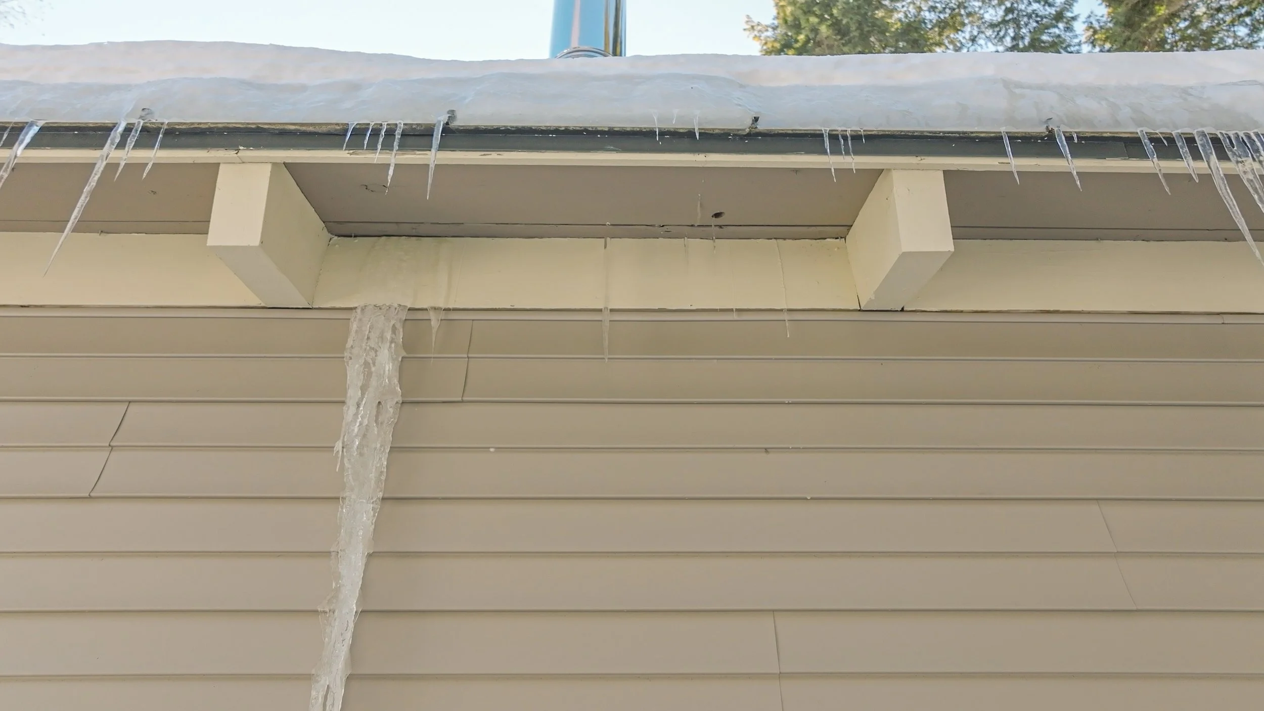 Icicles hanging from the edge of a roof with snow on top, and beige siding on a house