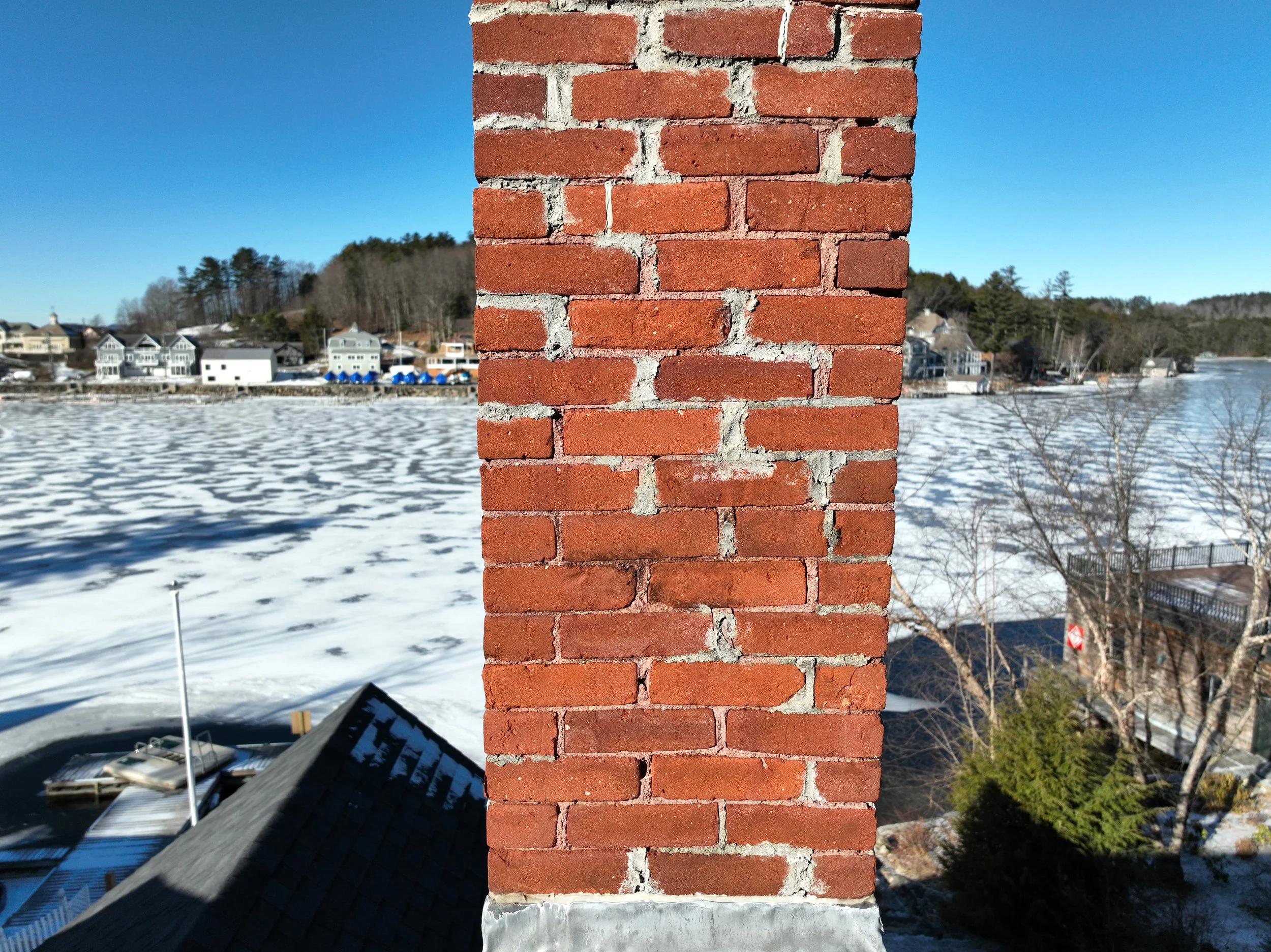 Close-up of a brick chimney on a house with a winter landscape in the background, including a partially frozen river, snow-covered ground, and houses along the shoreline under a clear blue sky.