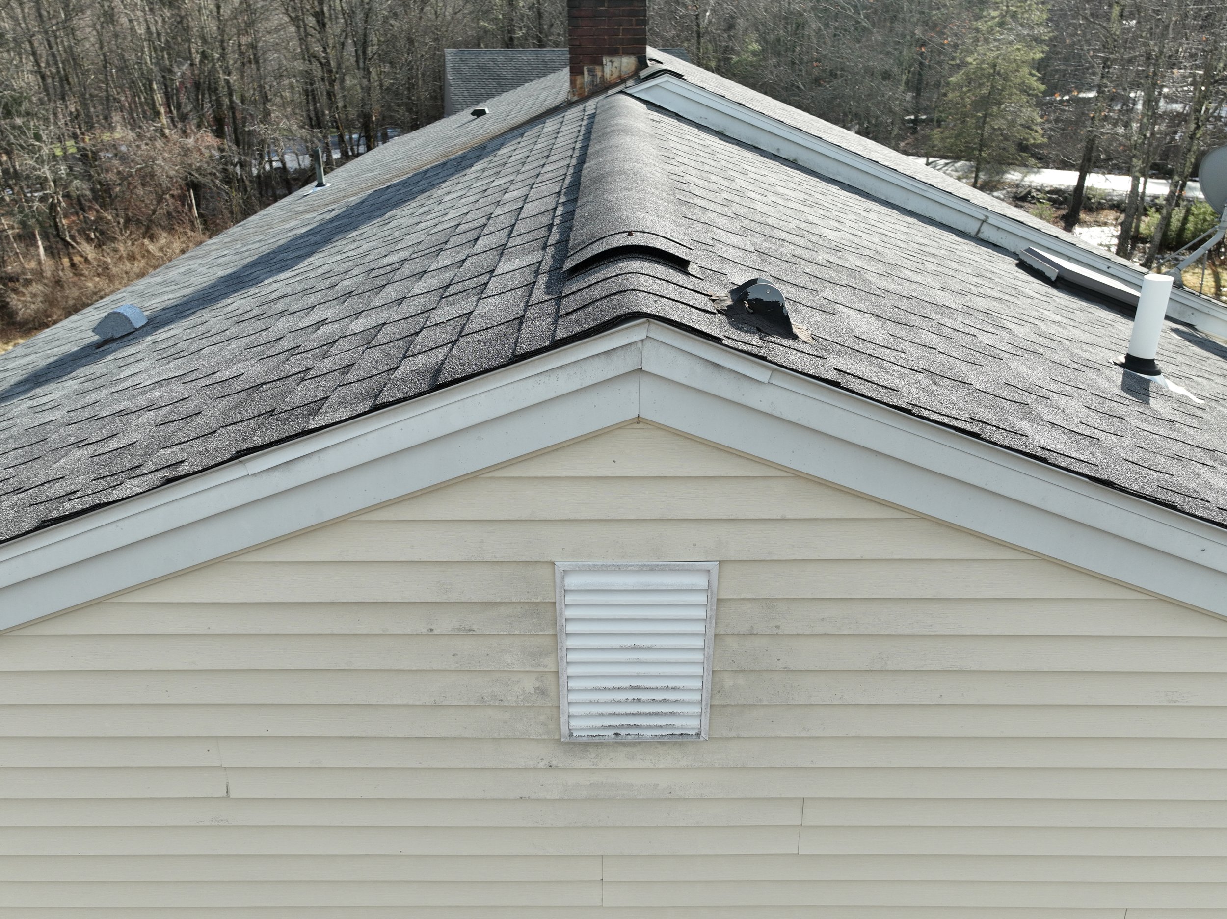 A house roof with shingles, a damaged section, and vents, with trees and other houses in the background.