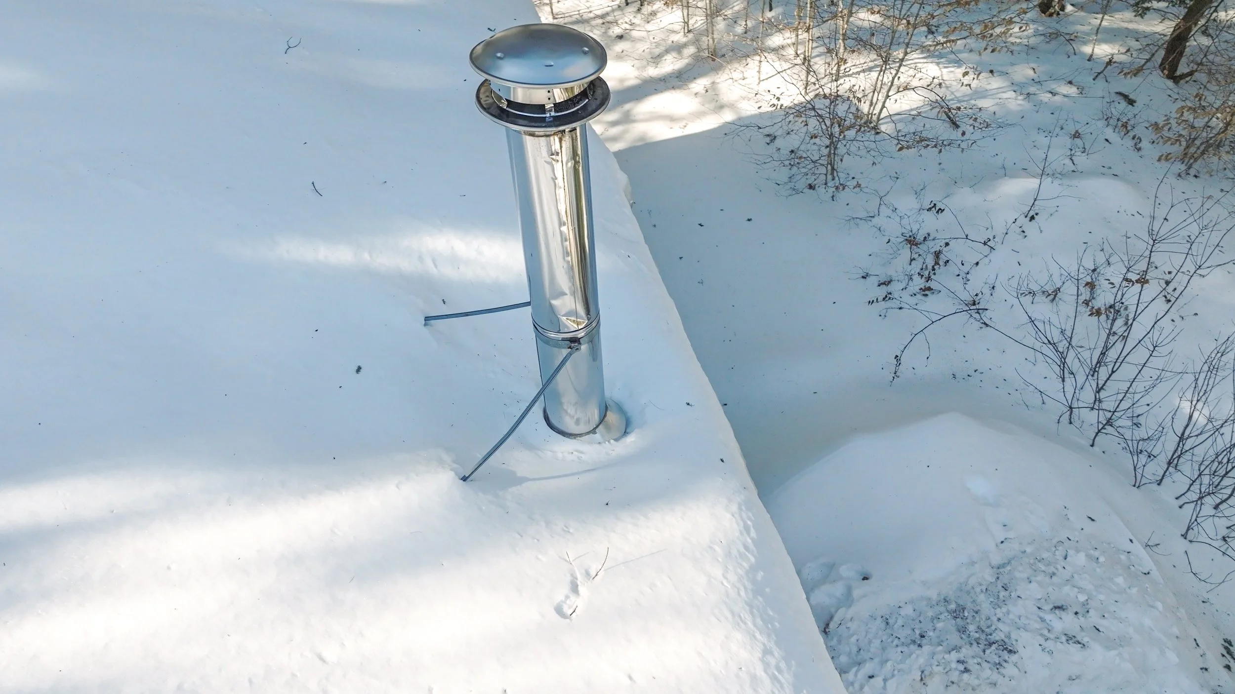Metal chimney vent pipe protruding from snow-covered ground with snow pile and leafless bushes in the background.