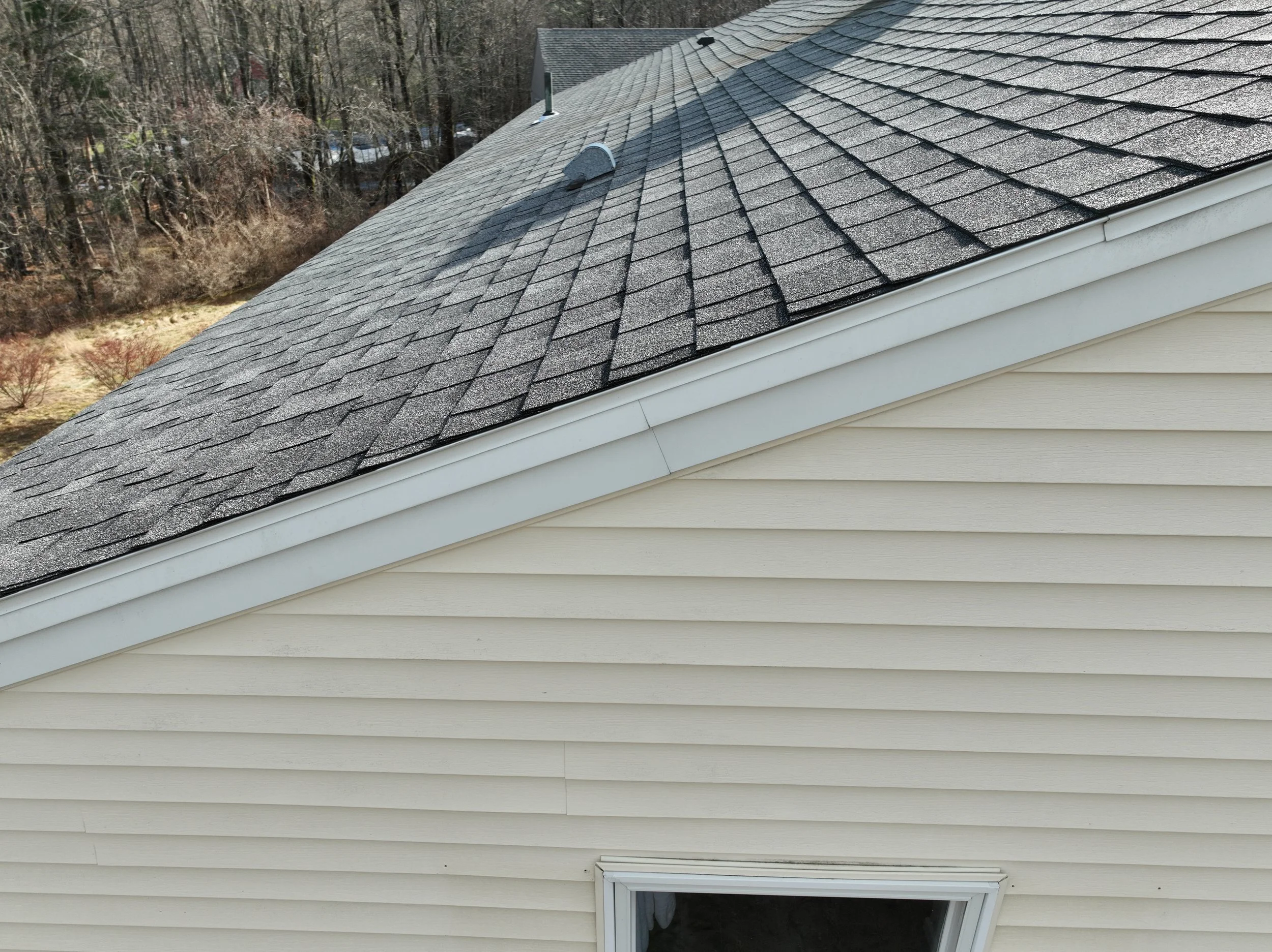 Close-up of a house's roof with black asphalt shingles and white vinyl siding, showing a window at the bottom.