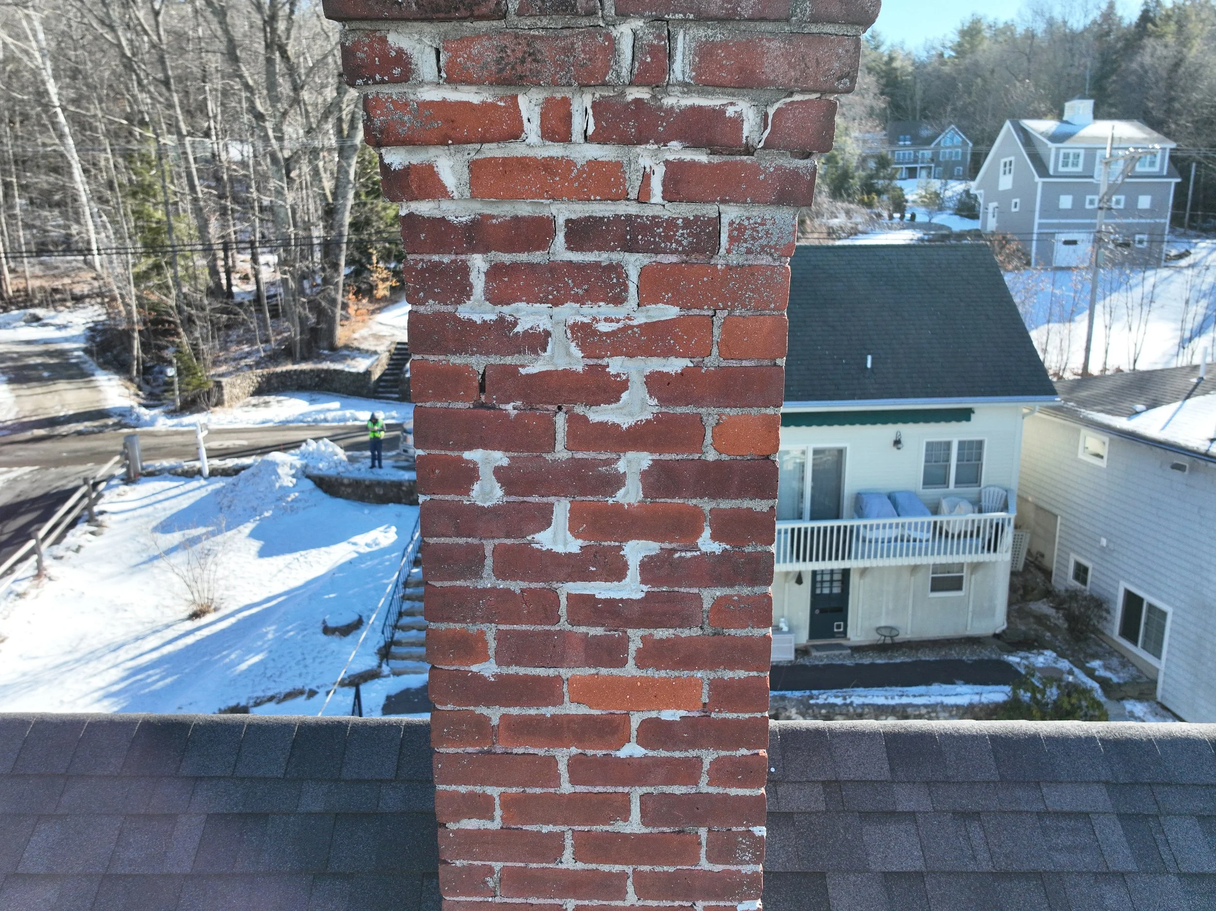 A red brick chimney on a house roof with snow-covered ground and neighboring houses in the background.