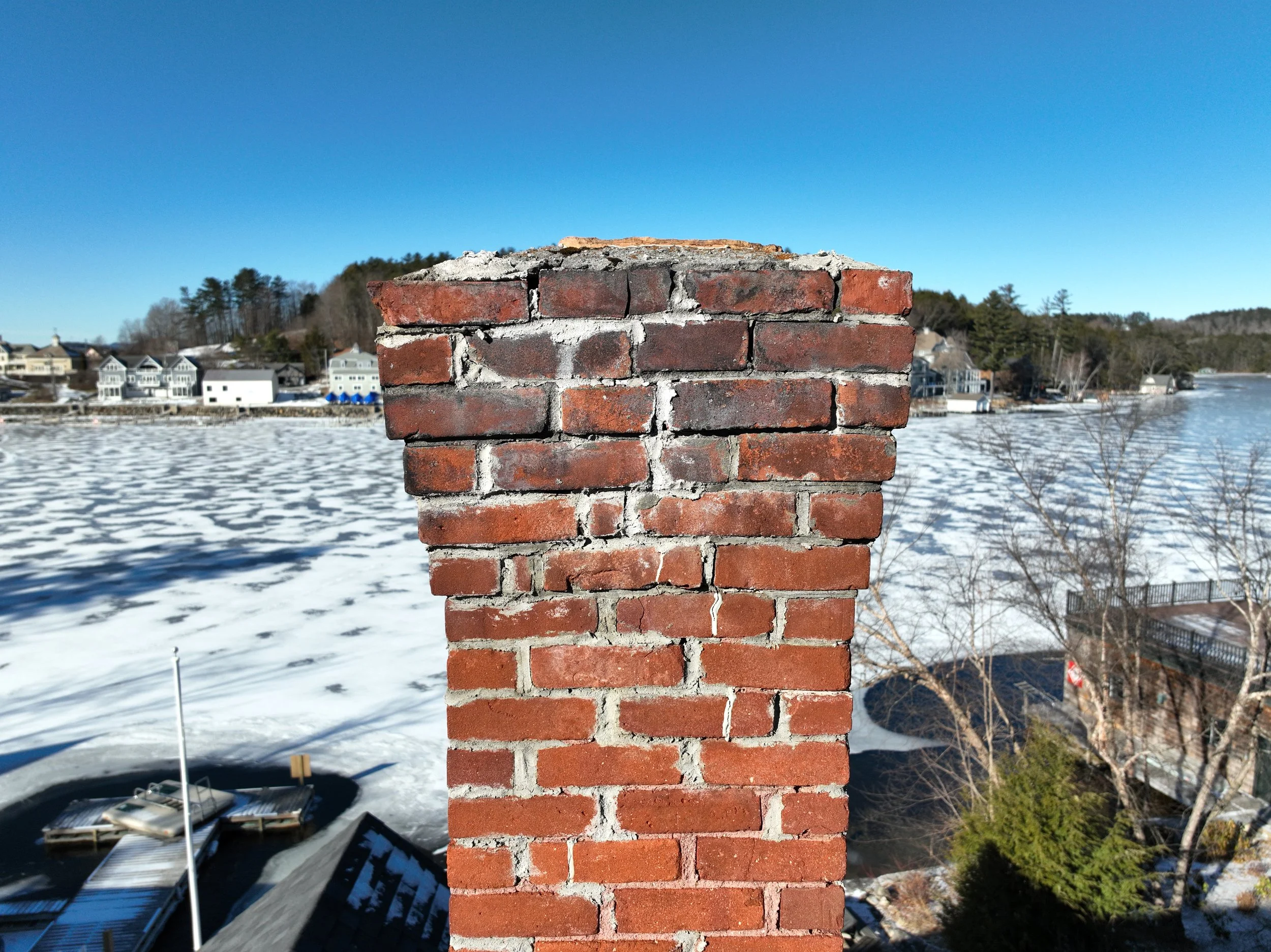 Close-up of a brick chimney in a winter landscape with a frozen body of water and shoreline houses in the background under a clear blue sky.