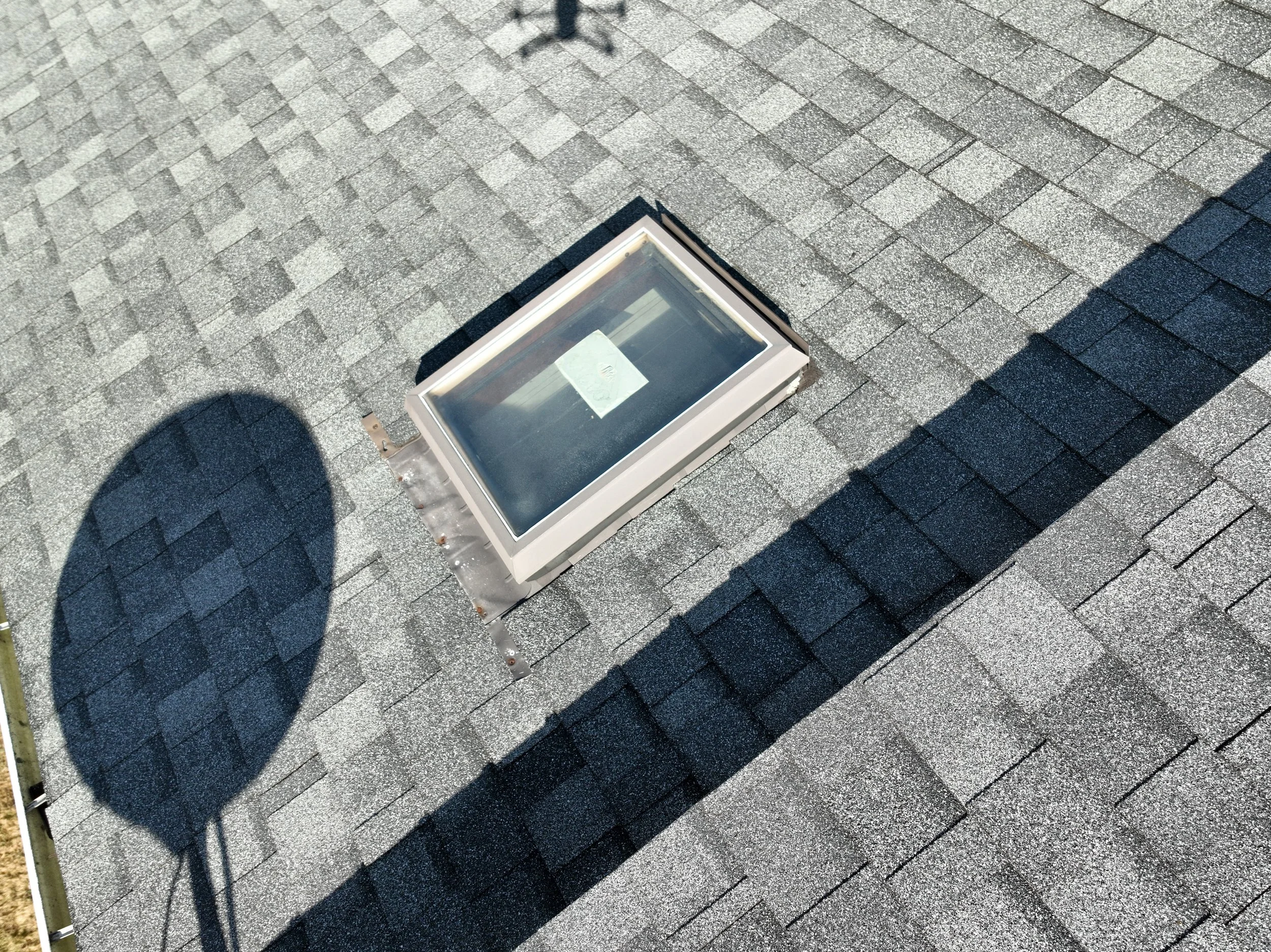 View of a roof with gray asphalt shingles, a skylight window, and the shadows of a chimney and a ladder cast on the shingles.