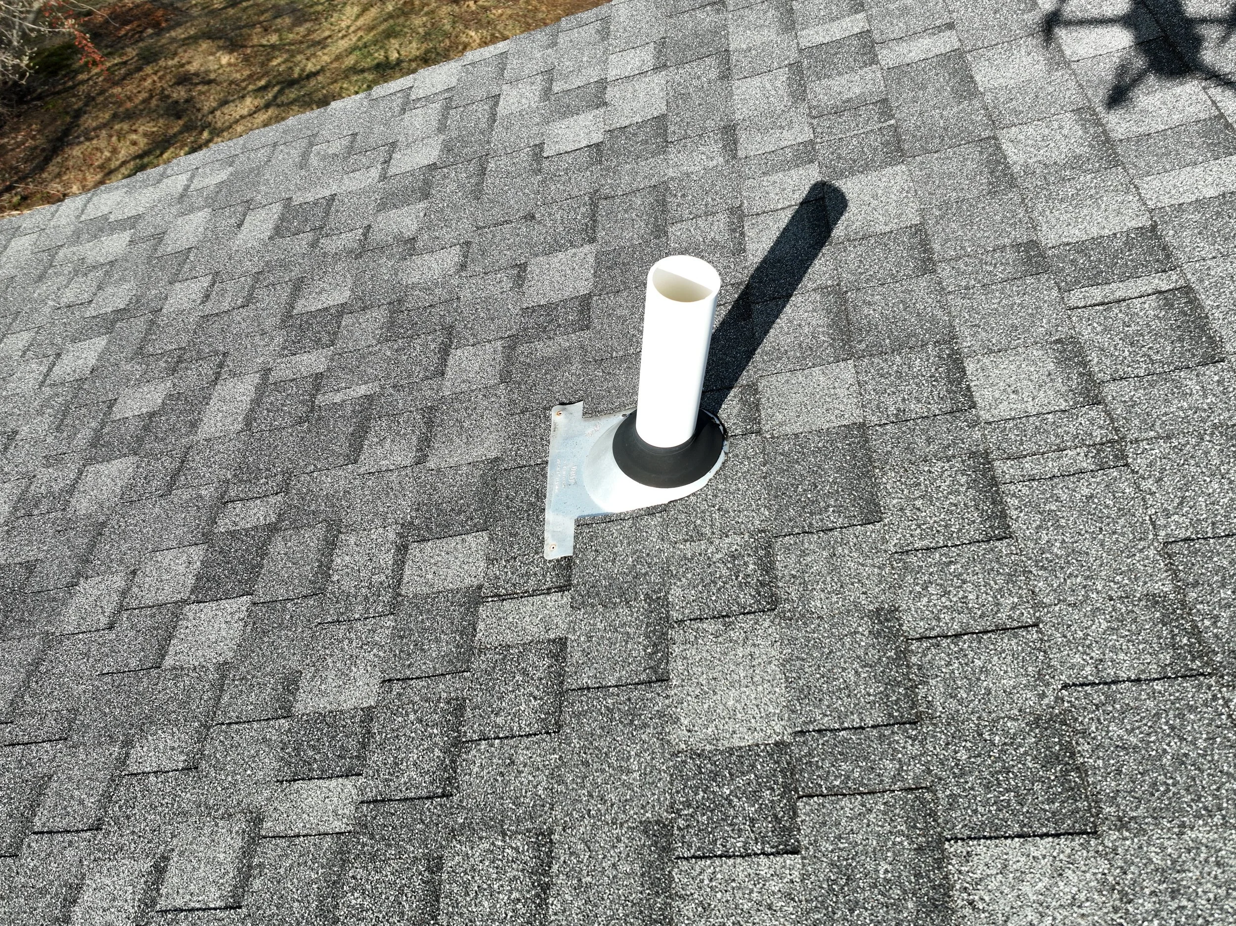 A vent pipe protruding from a shingled roof, casting a shadow to the right.