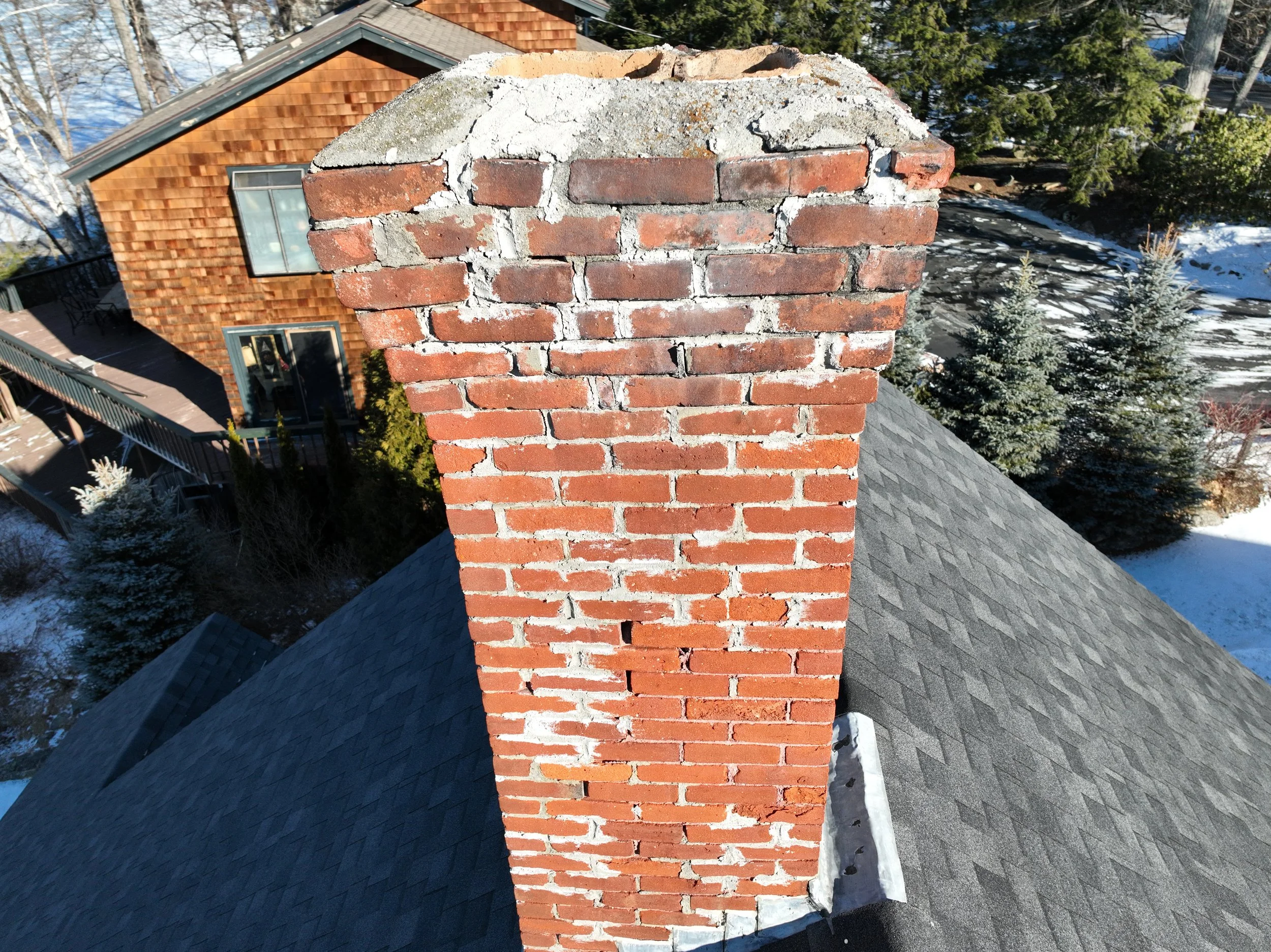 Close-up view of a brick chimney on a house rooftop, with trees and other houses in the background.