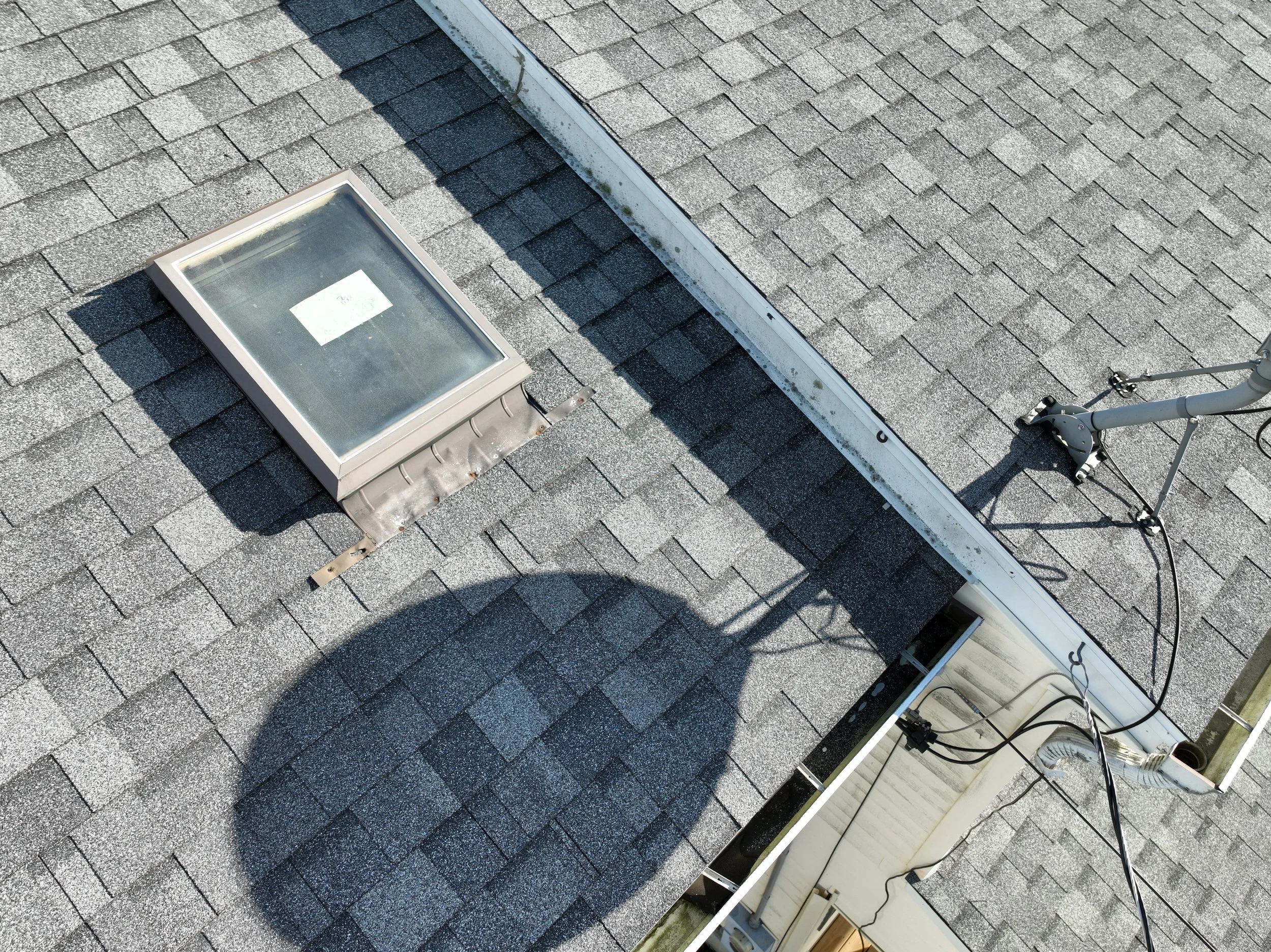 A rooftop with gray asphalt shingles, a roof vent with a glass cover, and an HVAC unit with exposed wiring and piping.