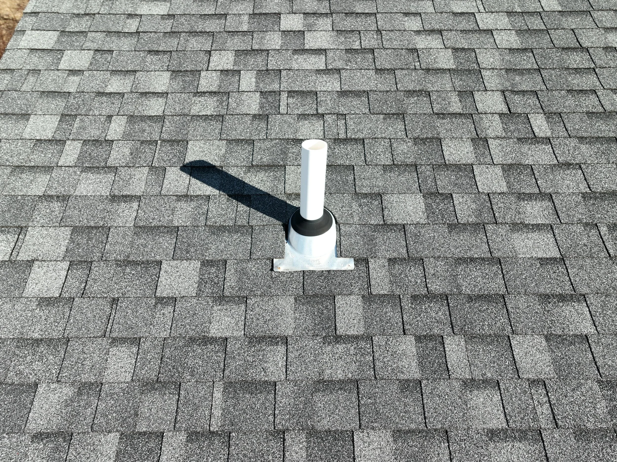Vent pipe on a shingled rooftop casting a shadow.