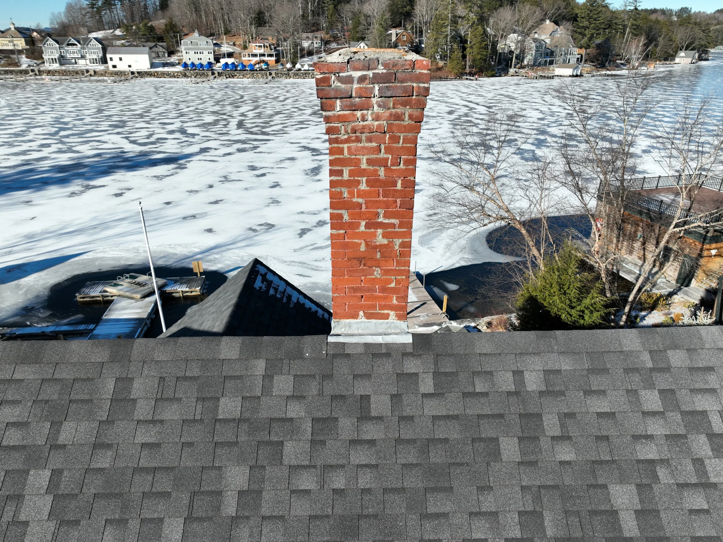 View of a snowy waterfront scene from a rooftop, featuring a brick chimney, rooftops of neighboring houses, frozen lake, and trees along the shoreline.