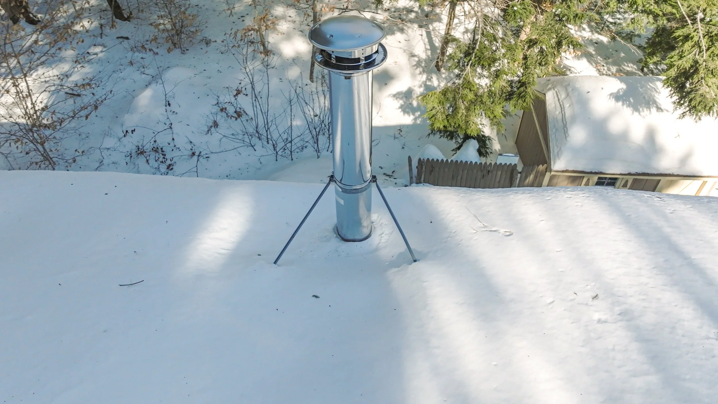 A metal chimney pipe on a snow-covered roof with trees and a shed in the background.