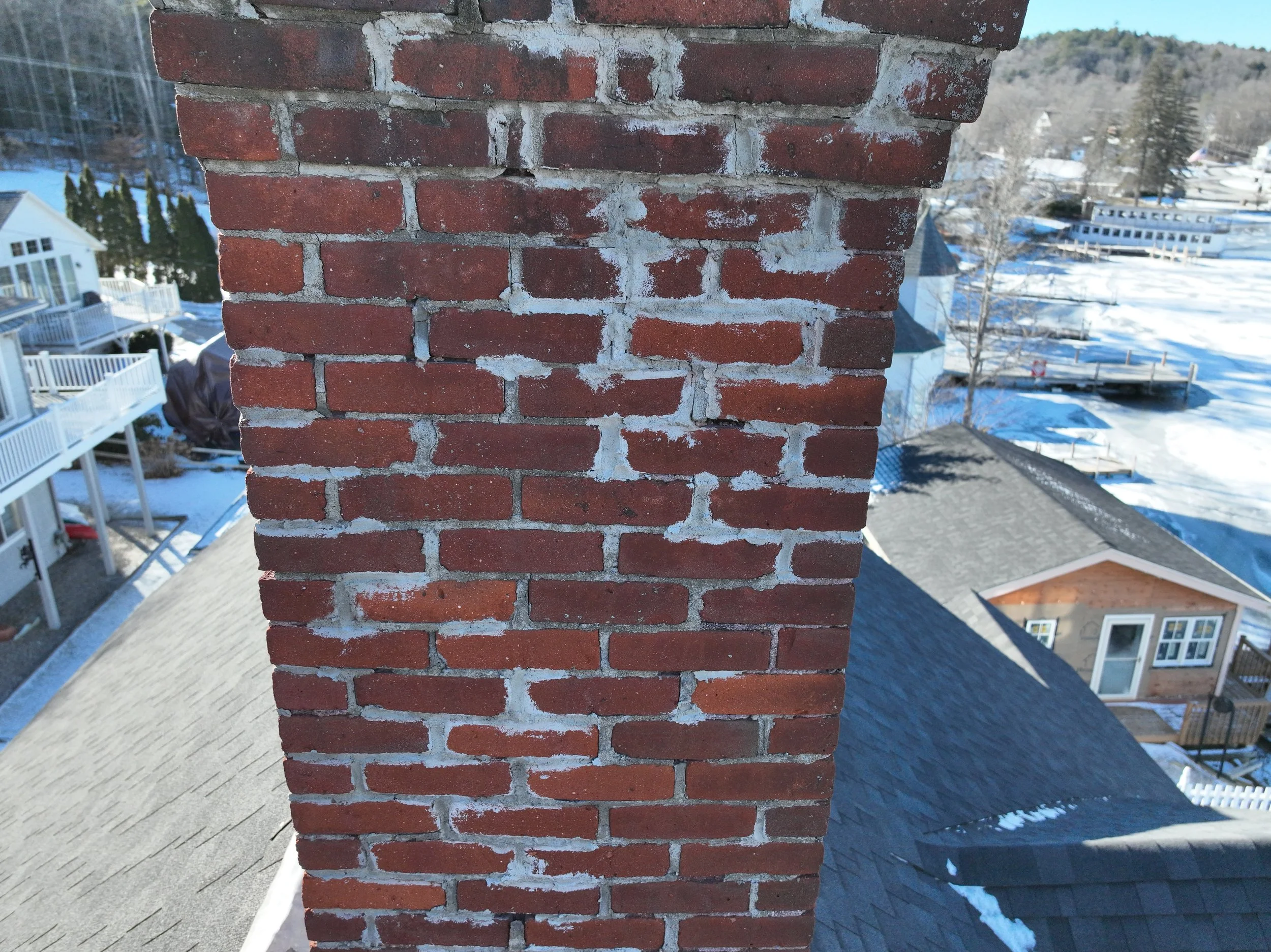 Close-up view of a brick chimney on the roof of a house, with snow visible on other rooftops and the ground in the background.
