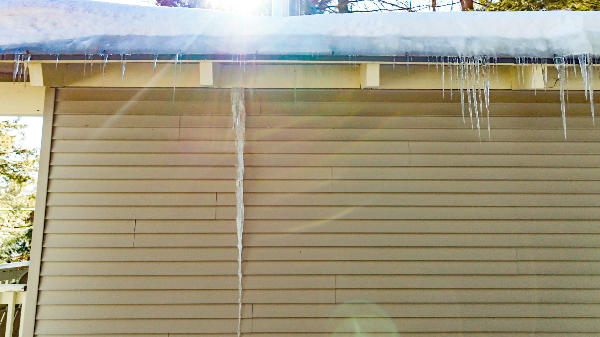 Icicles hanging from snow-covered roof above beige siding. Sunlight creating lens flare.