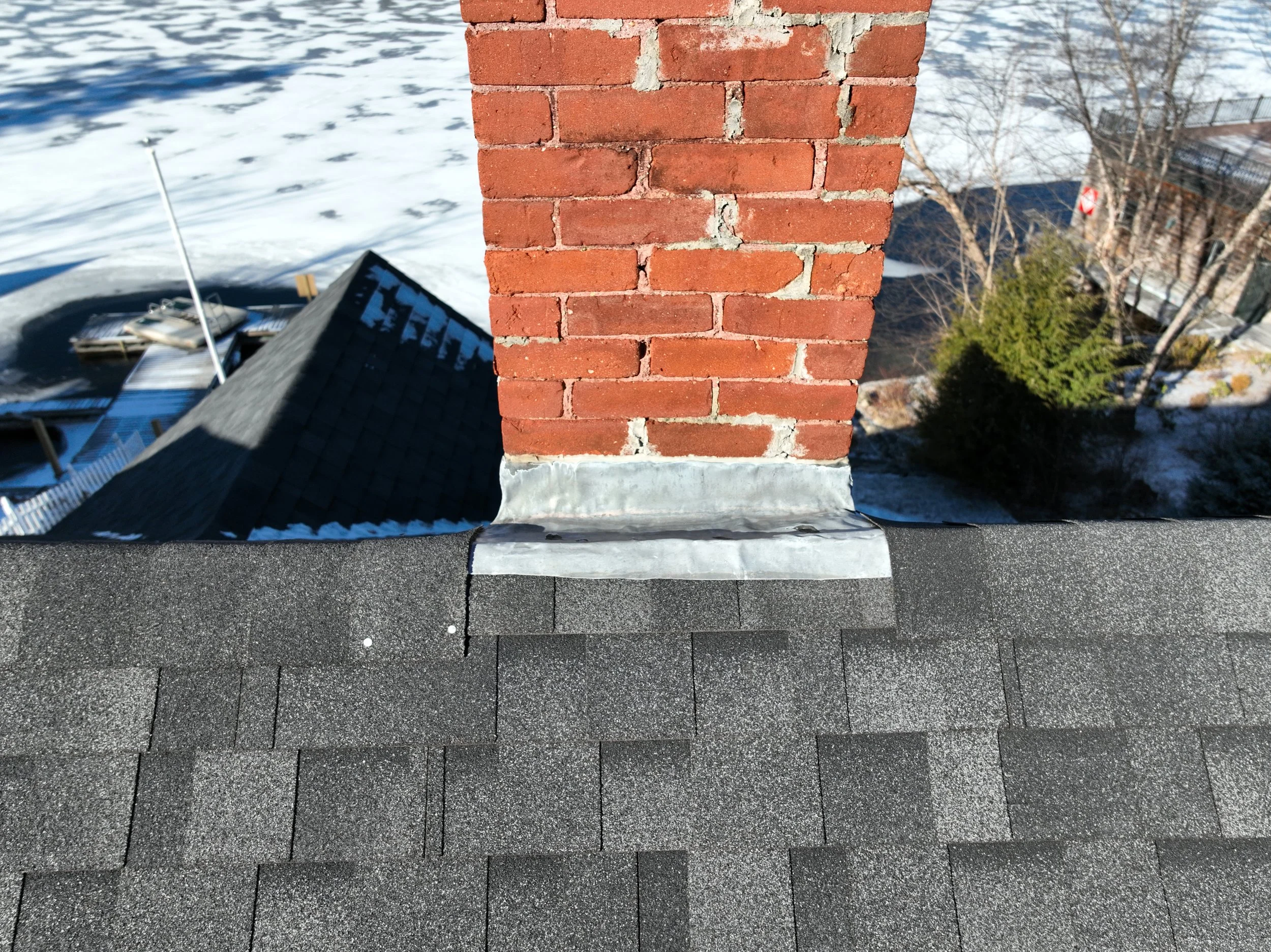 View from a rooftop showing a brick chimney with flashing, asphalt shingles, and neighboring rooftops with snow-covered yards, trees, and buildings.