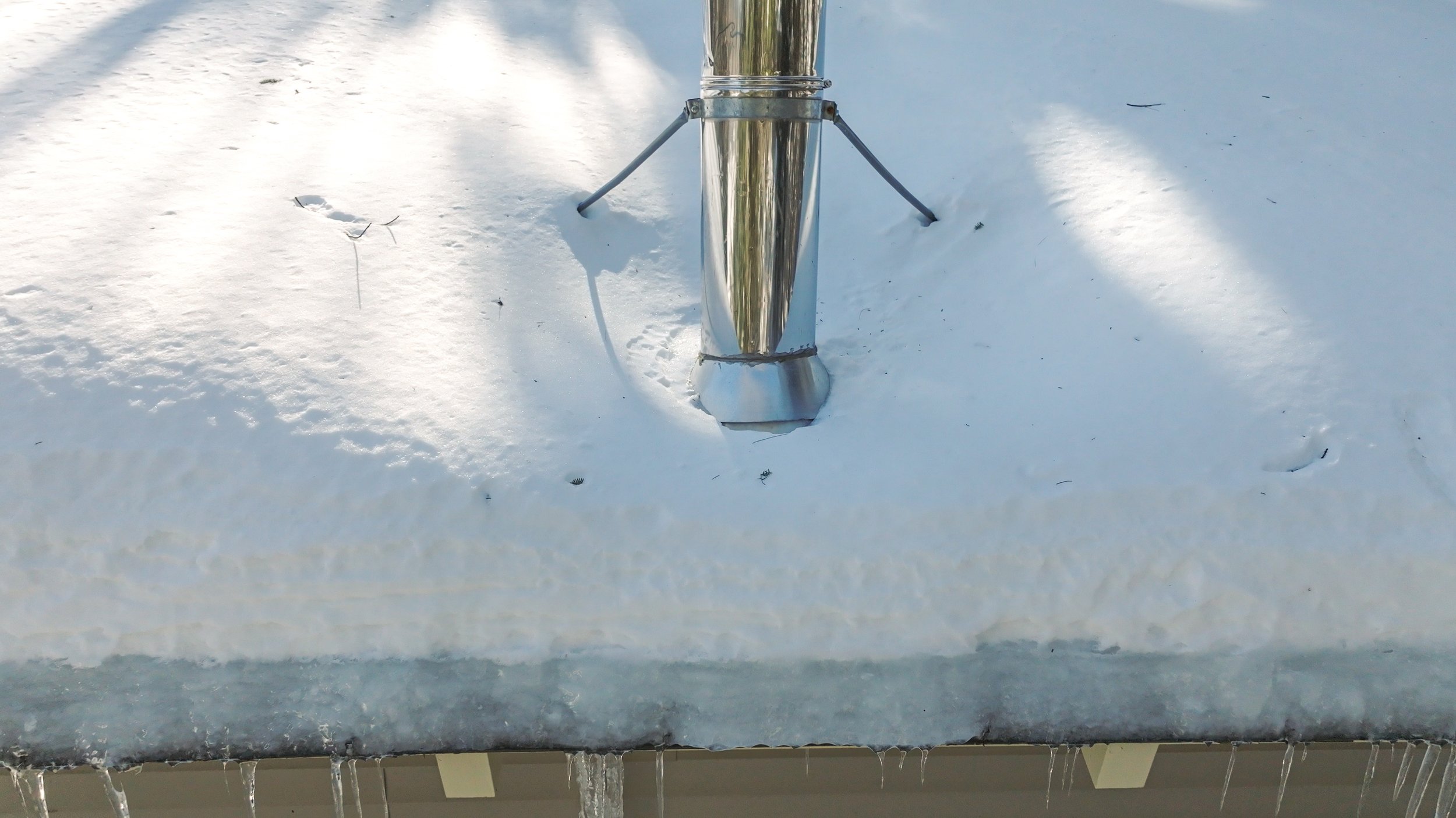 Close-up of a metal pole on a snowy rooftop, with snow and ice surrounding it and icicles hanging from the edge.