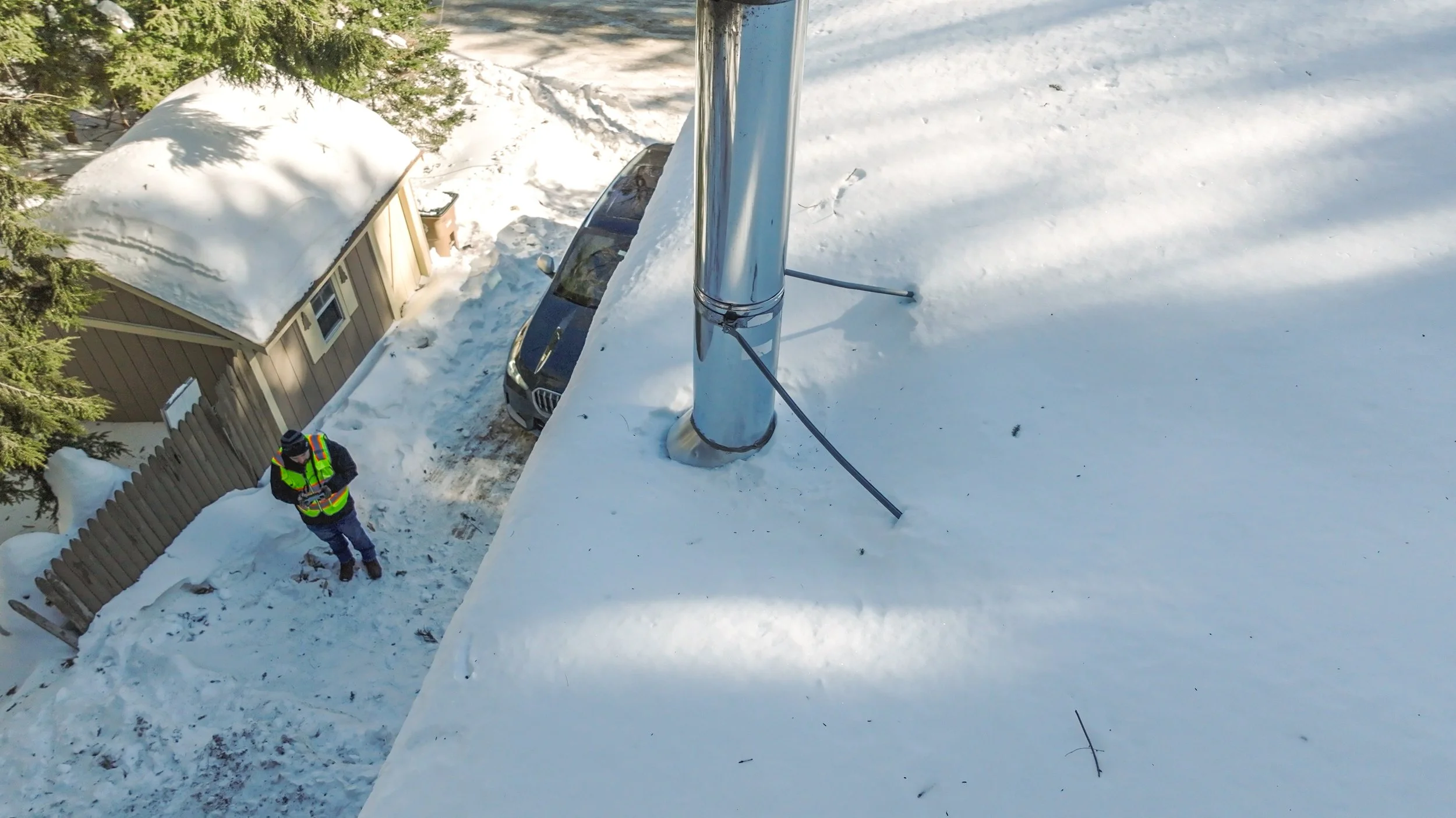 A person wearing a flashing safety vest standing on snow-covered ground near a wooden fence and a small house, taken from a high vantage point.
