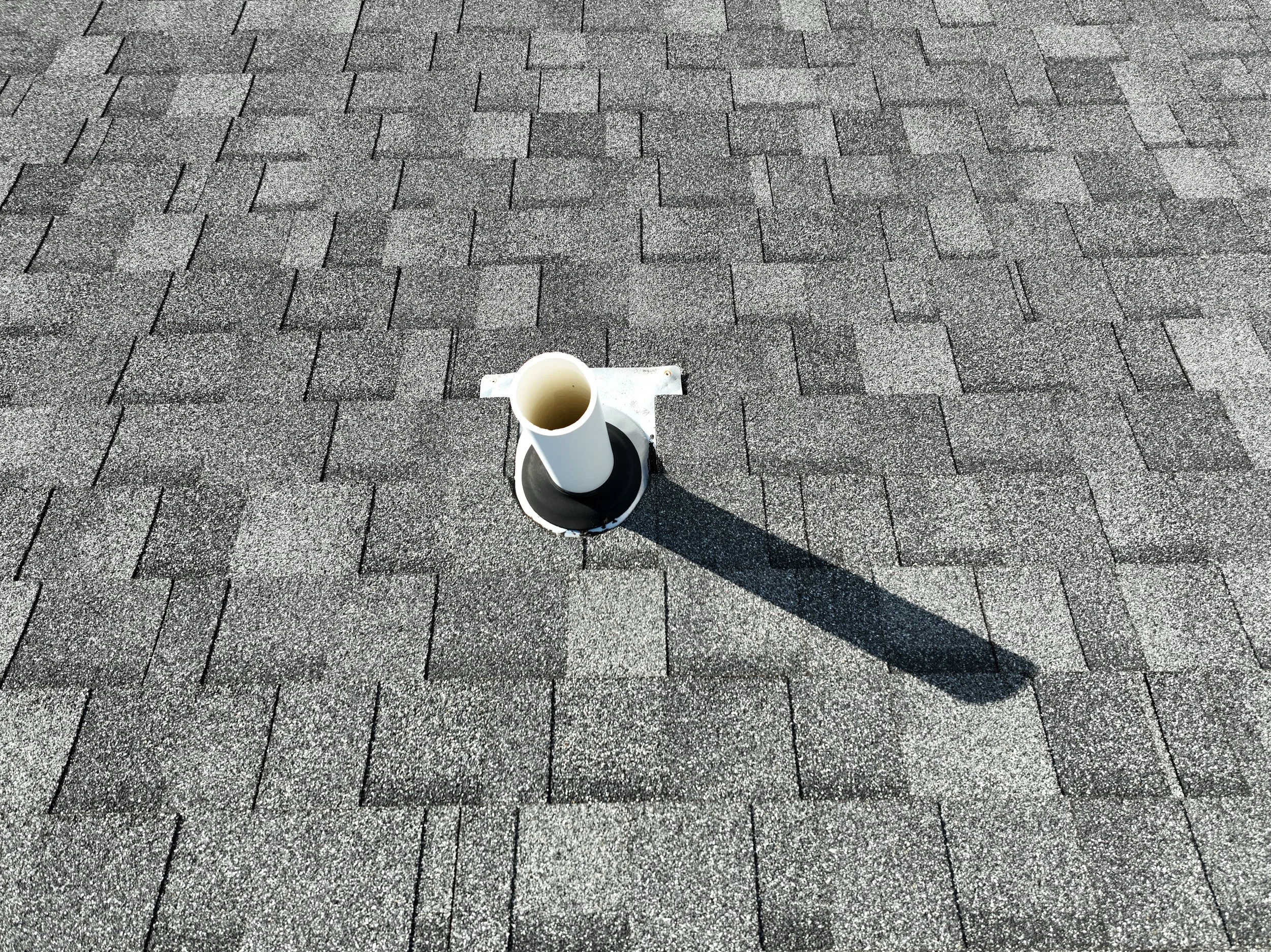 A white vent pipe protruding from the gray asphalt shingled roof with a shadow cast to the right.