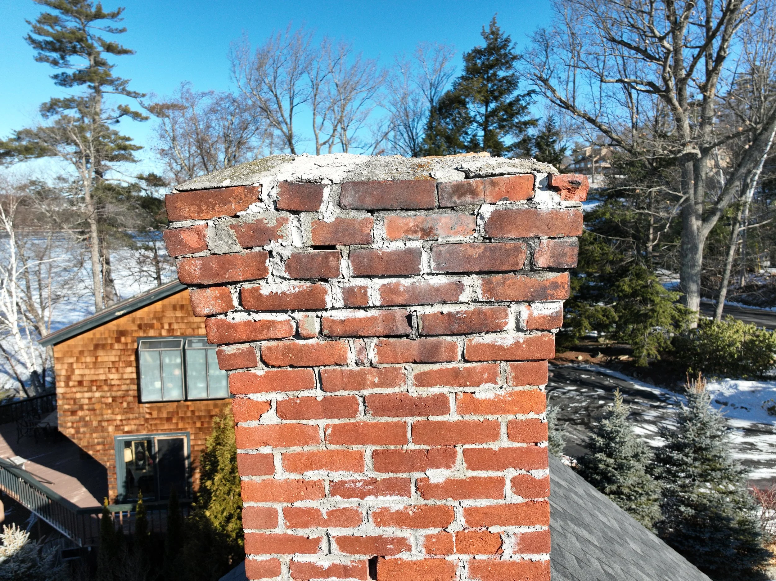 Close-up of a brick chimney on the roof of a house, with a background of trees and a clear blue sky.
