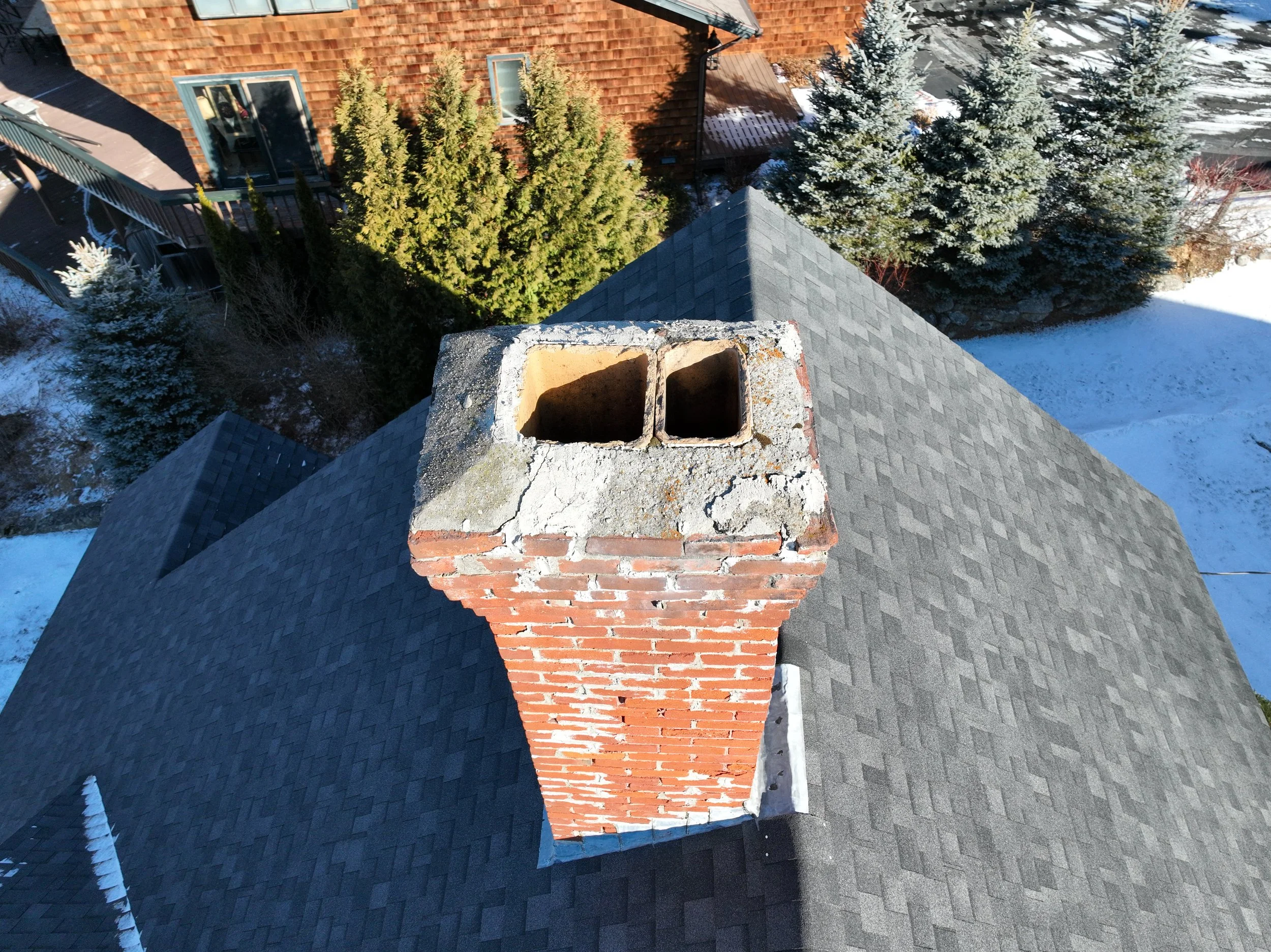 View from the top of a house's roof showing a brick chimney with two hollow rectangular openings. Snow covers parts of the roof and surrounding trees, with a neighboring house and trees in the background.