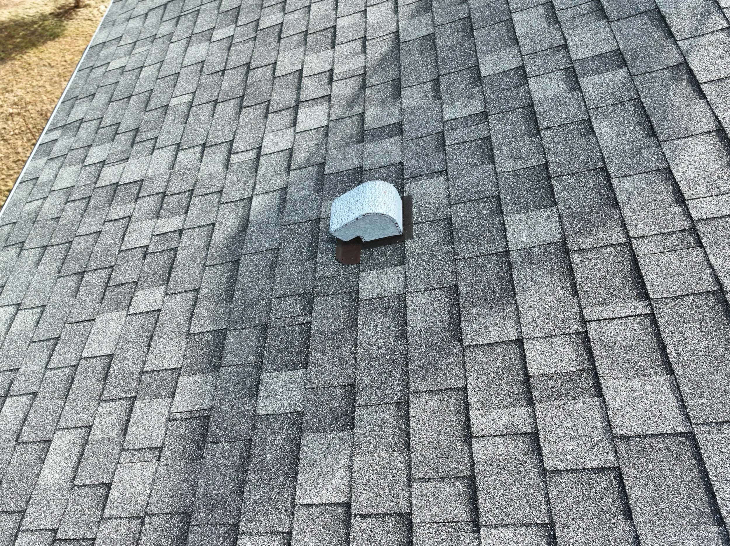 Close-up of a shingled roof with a small white vent or cover and dark metal flashing.