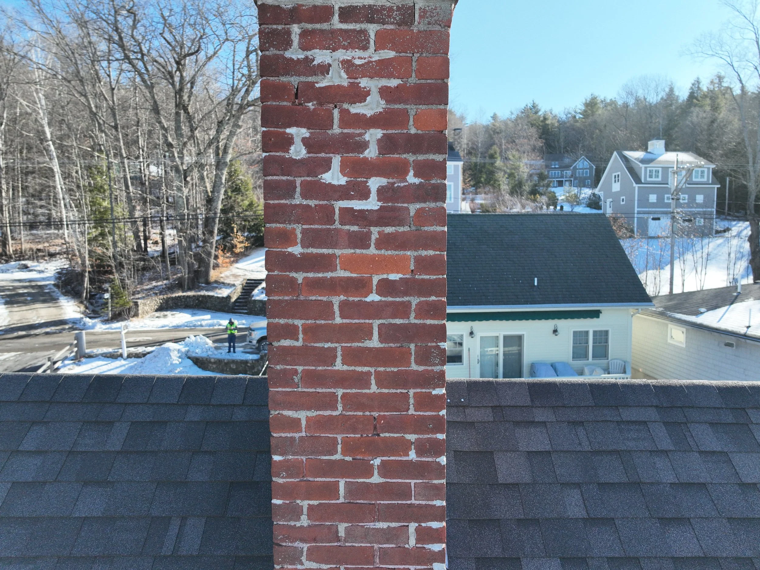 View of rooftops and houses from a window with a central brick chimney in the foreground, snow on the ground, leafless trees, and a person in a reflective vest near a car in a residential neighborhood.
