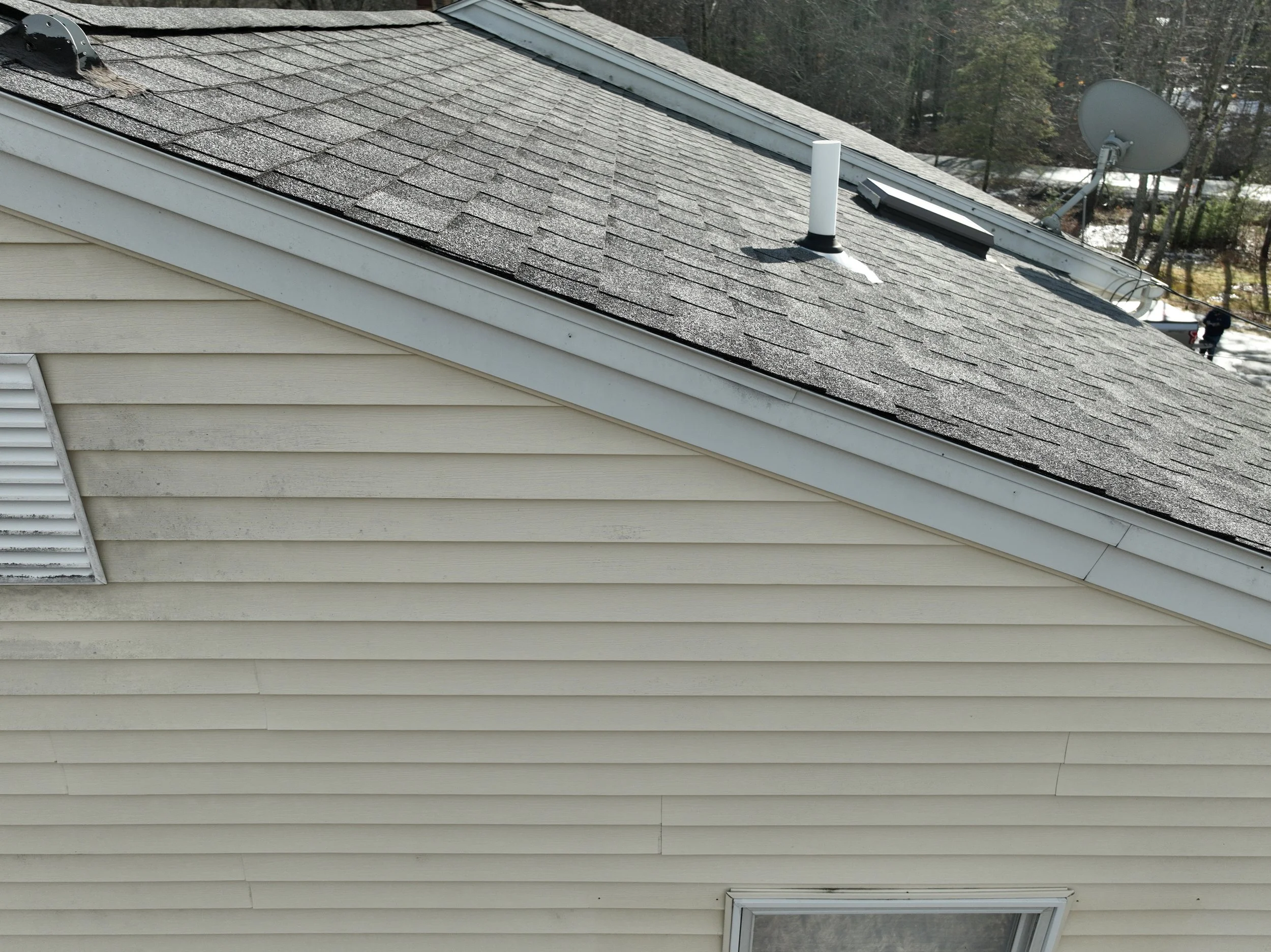 Close-up view of a house's beige vinyl siding exterior, including a window and a section of the roof with asphalt shingles, a vent pipe, and a satellite dish.