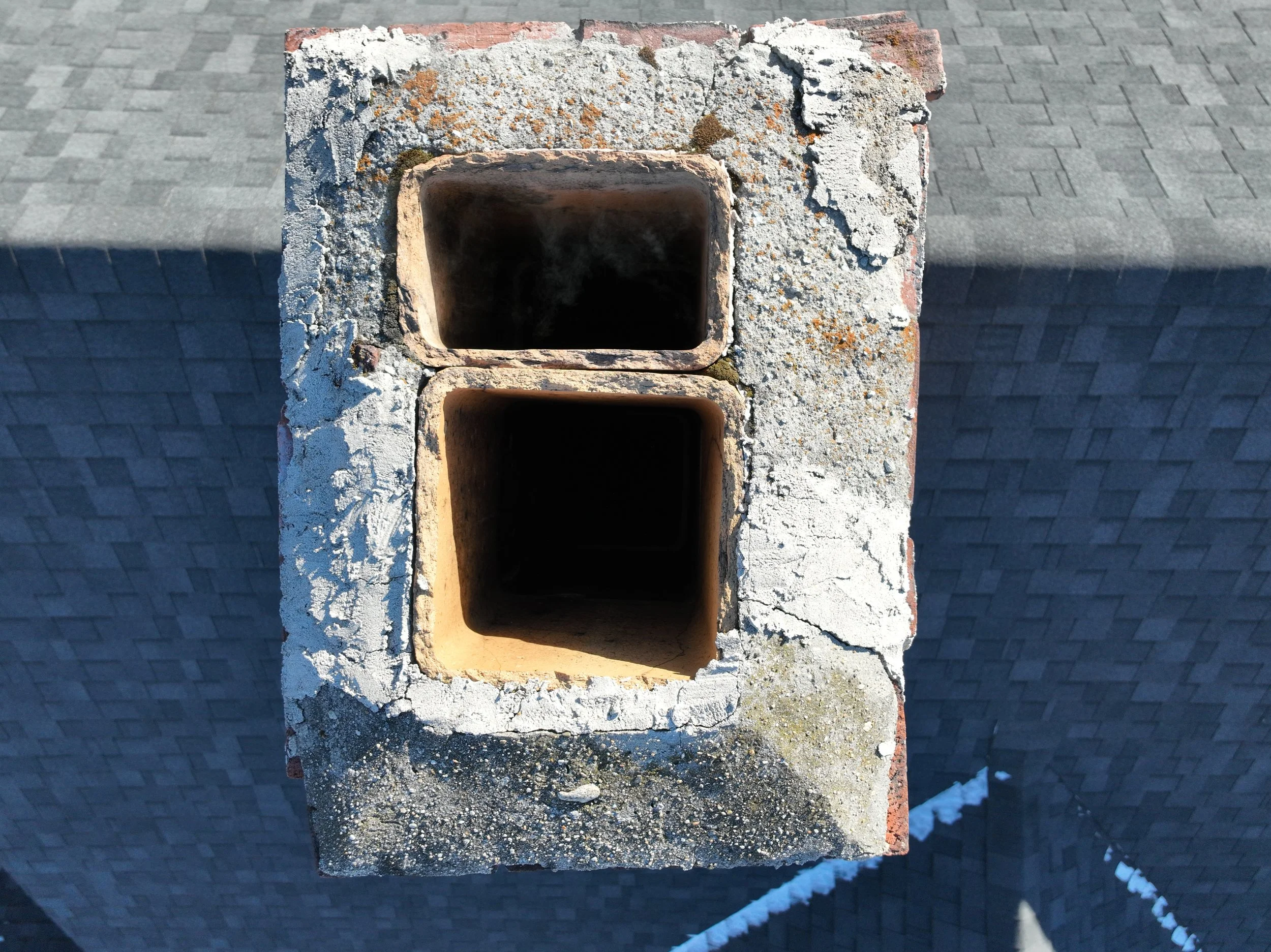 Top view of a brick chimney with two rectangular flues, one above the other, on a rooftop with gray shingles.
