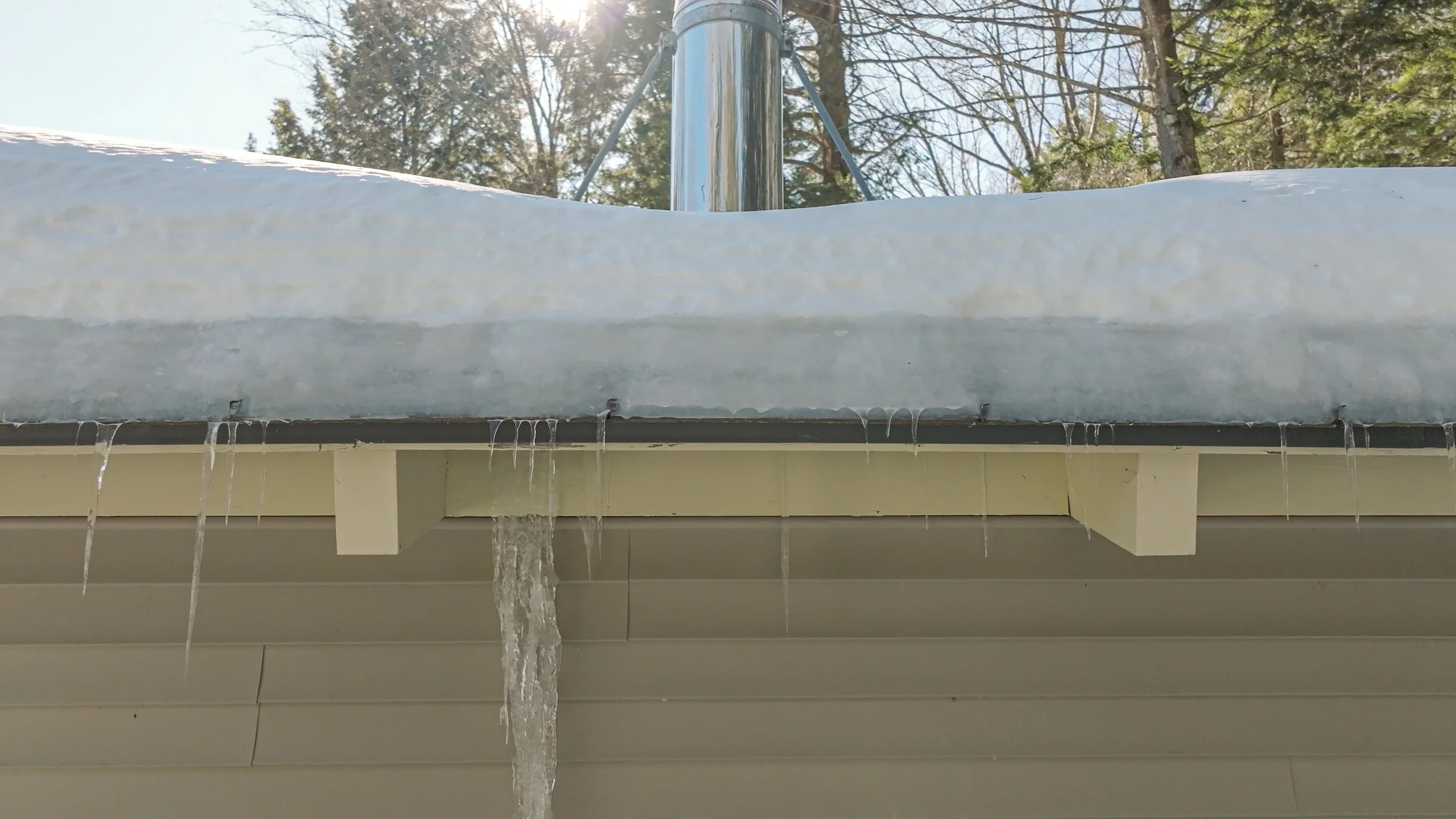 Icicles hanging from the edge of a snow-covered roof with a water stream dripping down.