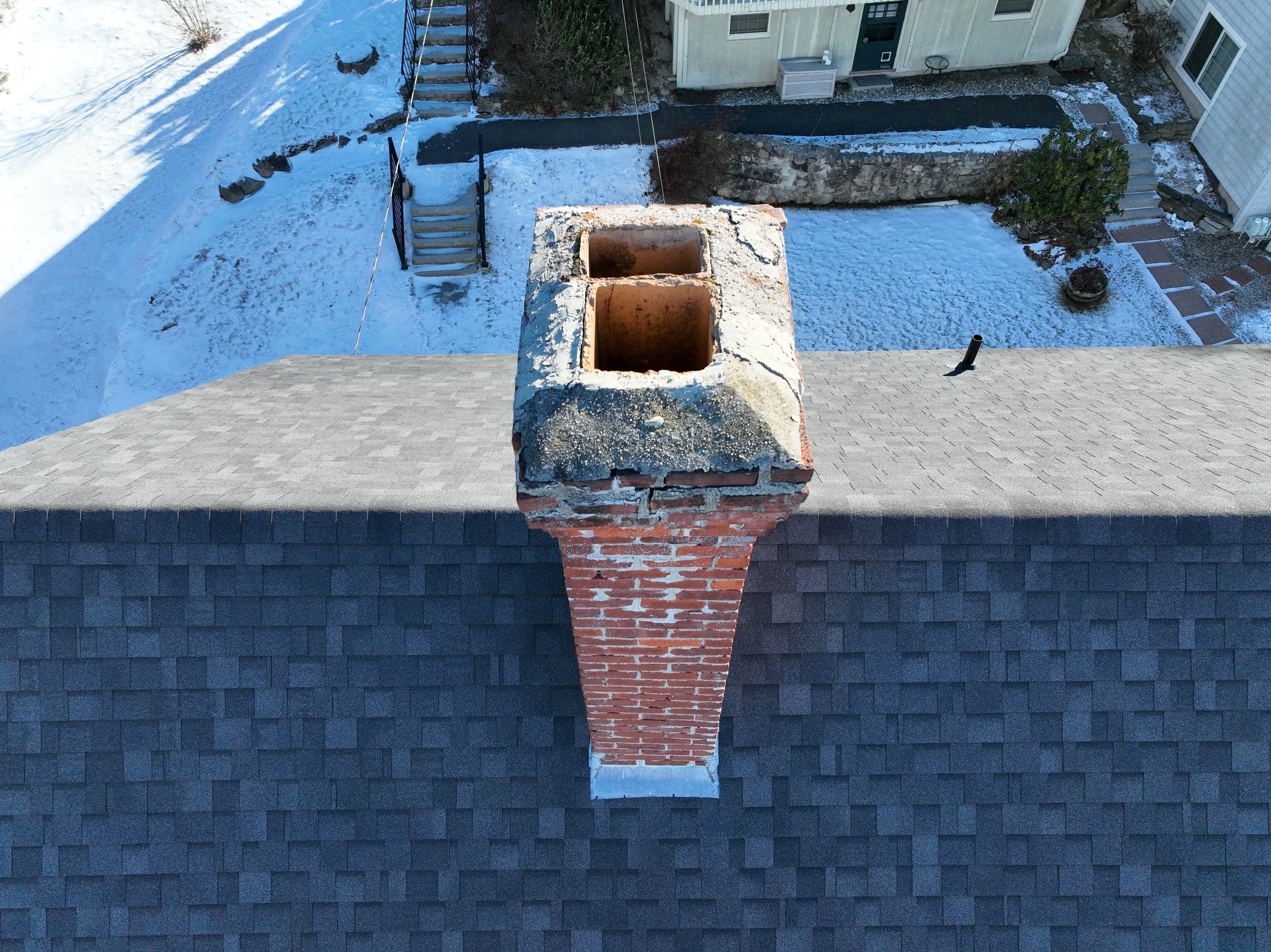 View of a rooftop chimney with two large metal flues, brick exterior, and a concrete cap, overlooking snow-covered yard with stairs, plants, and neighboring houses.