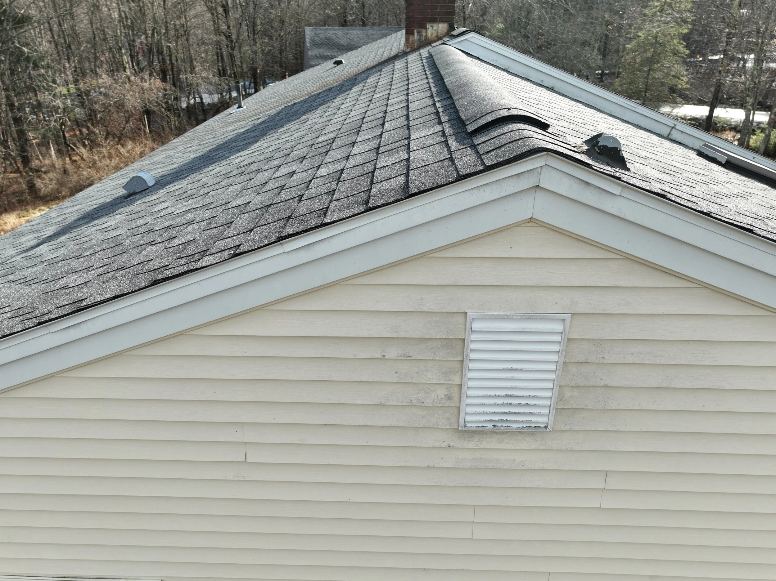 Close-up view of a house rooftop with asphalt shingles, a storm vent, and a ridgeline vent, with weathered beige siding on the house wall.