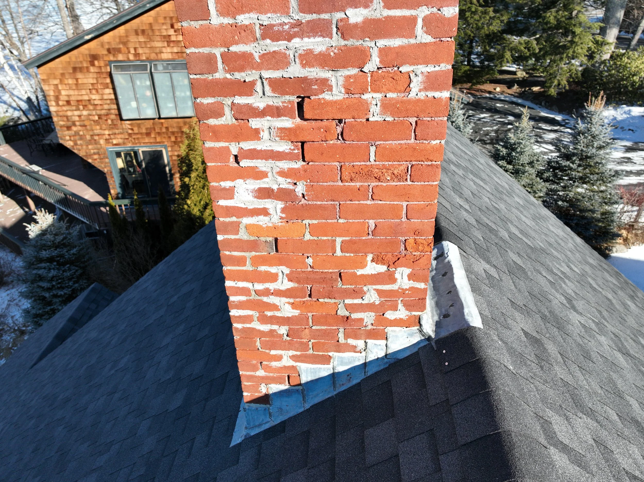 Close-up view of a brick chimney rising above a gray shingled roof of a house, with neighboring houses and snow-covered trees in the background.
