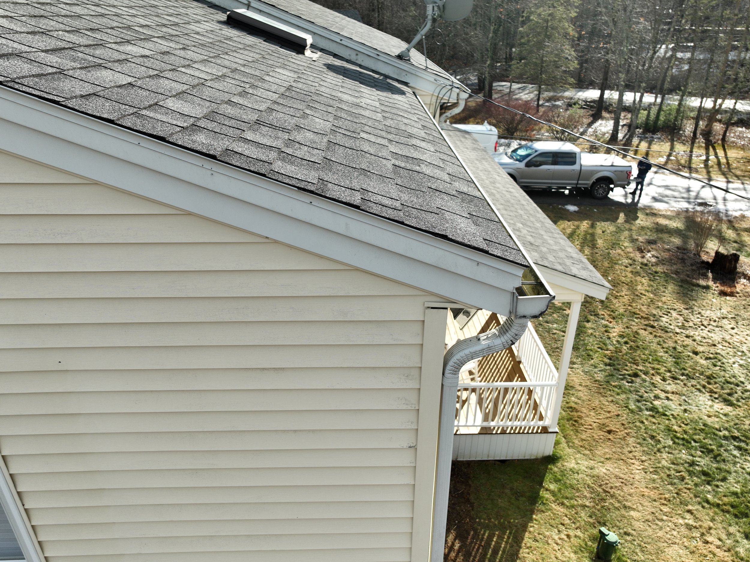A beige house with vinyl siding and a grey shingled roof, showing a gutter system. In the background, a grey pickup truck is parked on a snowy driveway with two people near it. The yard has grass and trees, some of which have no leaves.