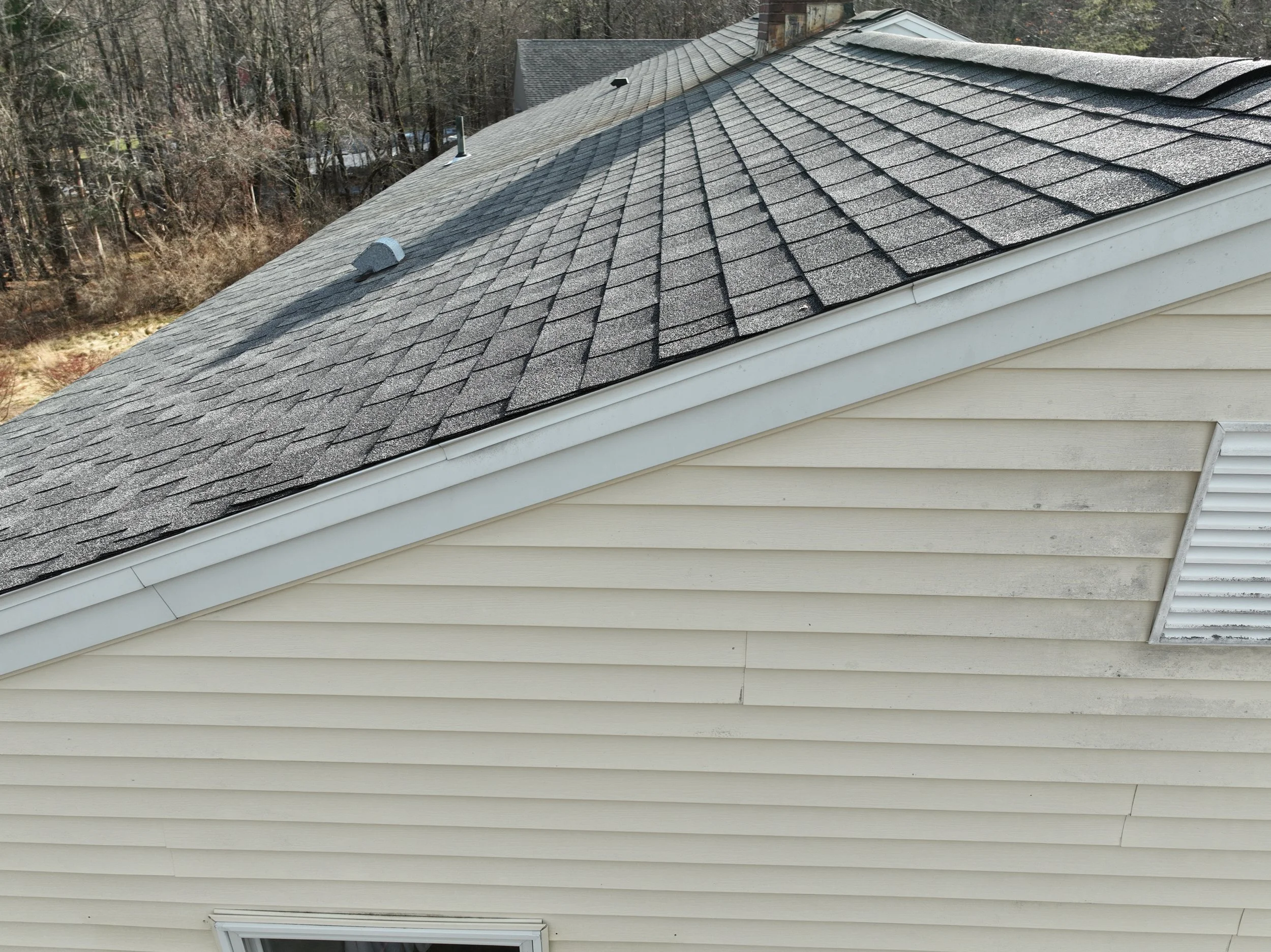 Close-up of a house roof with asphalt shingles and part of the house siding with horizontal beige vinyl siding.