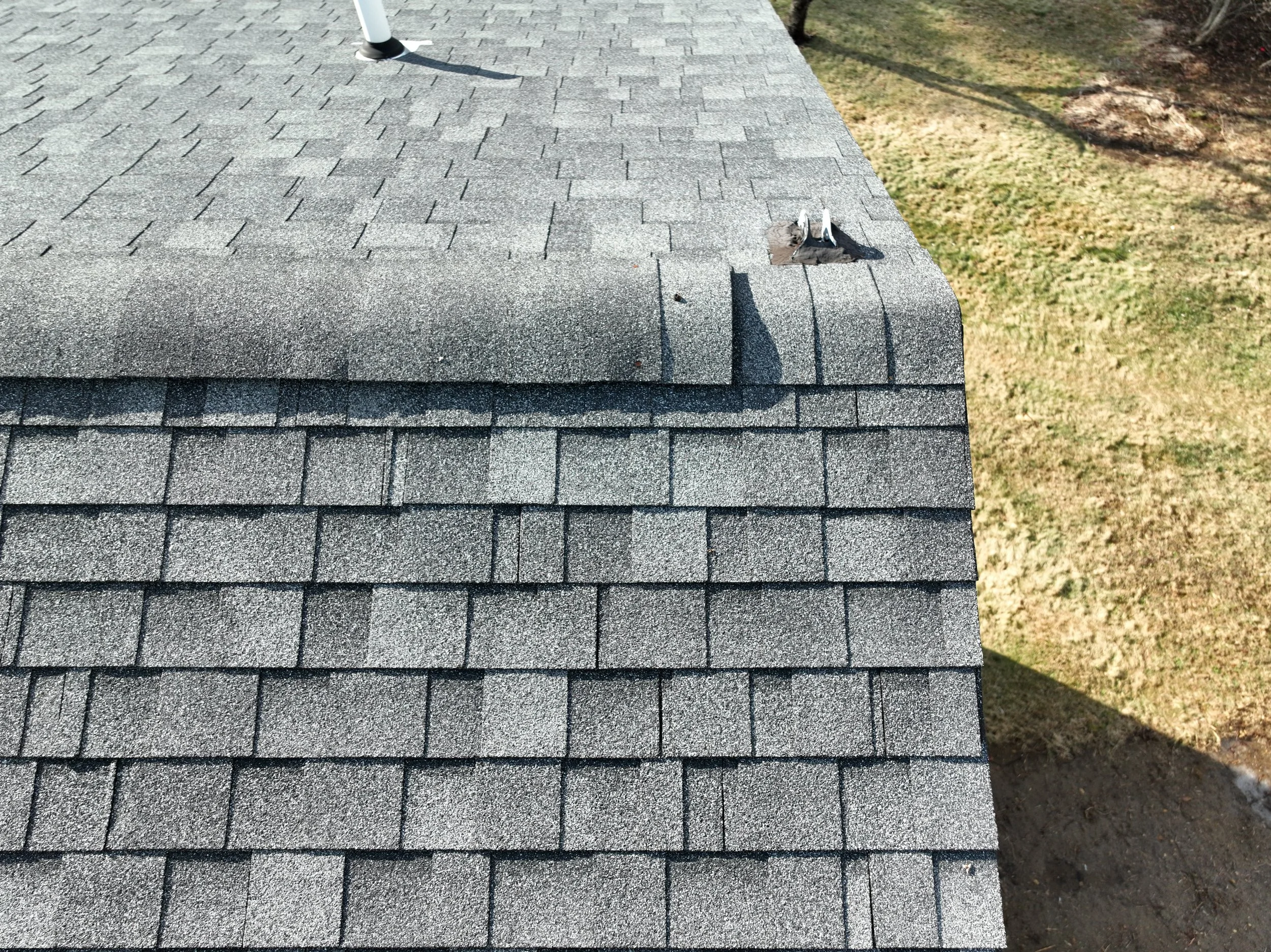 Close-up of a roof with gray asphalt shingles, showing a section with a missing shingle and some debris on the roof, with a grassy yard in the background.