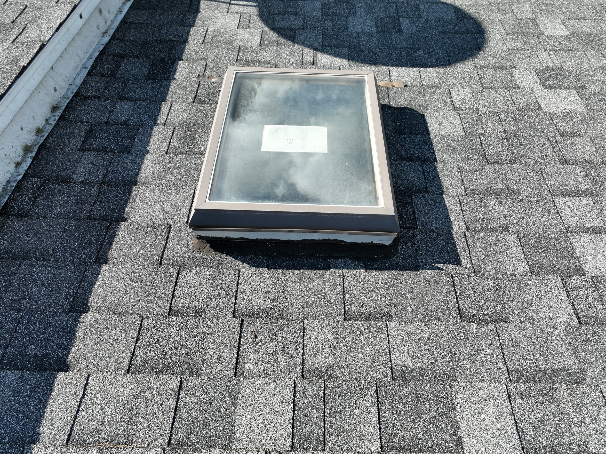 A skylight window on a shingled rooftop reflecting the sky and clouds.