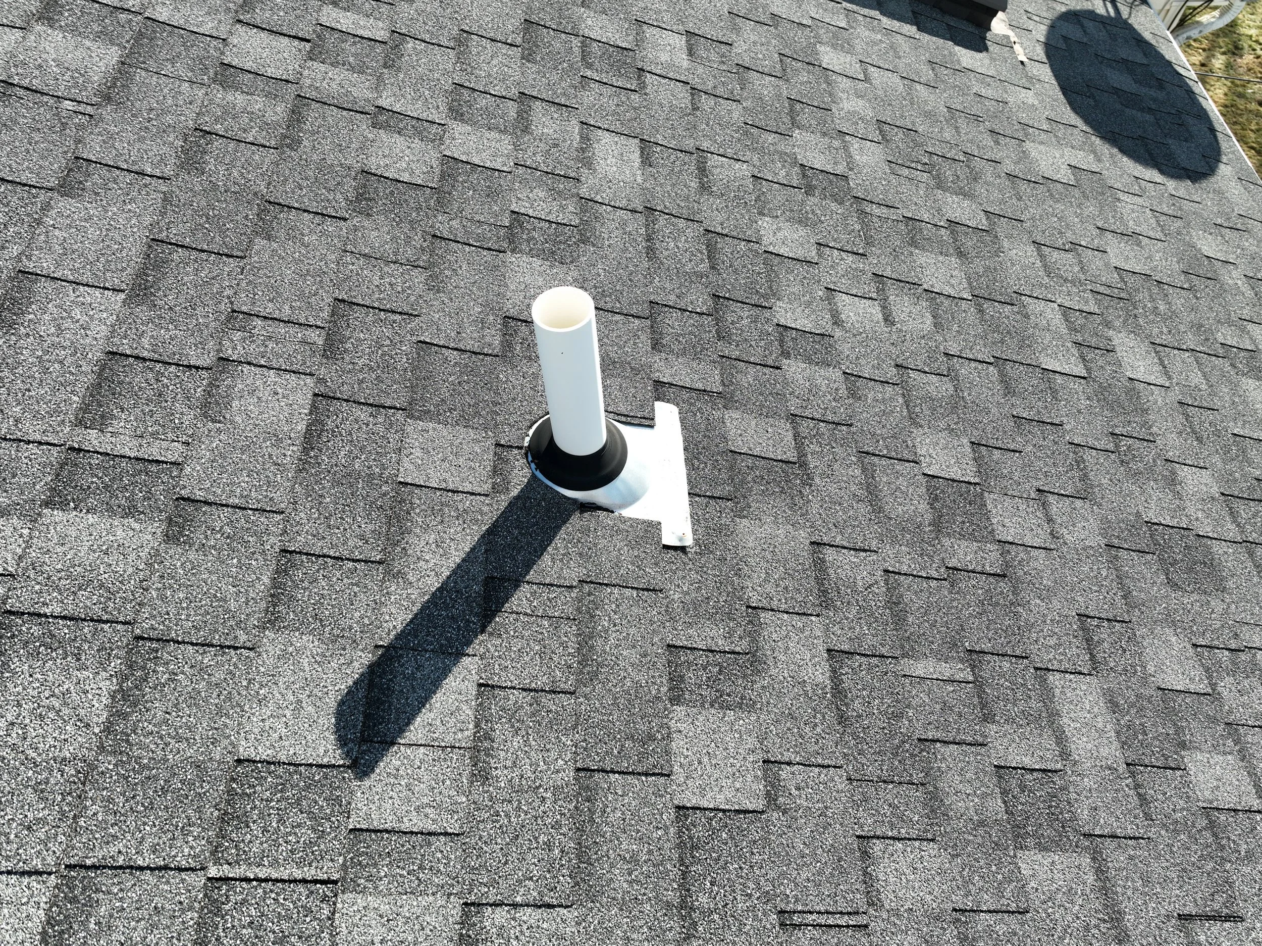 Roof with asphalt shingles and a white vent pipe with a black collar mounted on a metal flashing. The shadow of the vent pipe is cast on the roof.