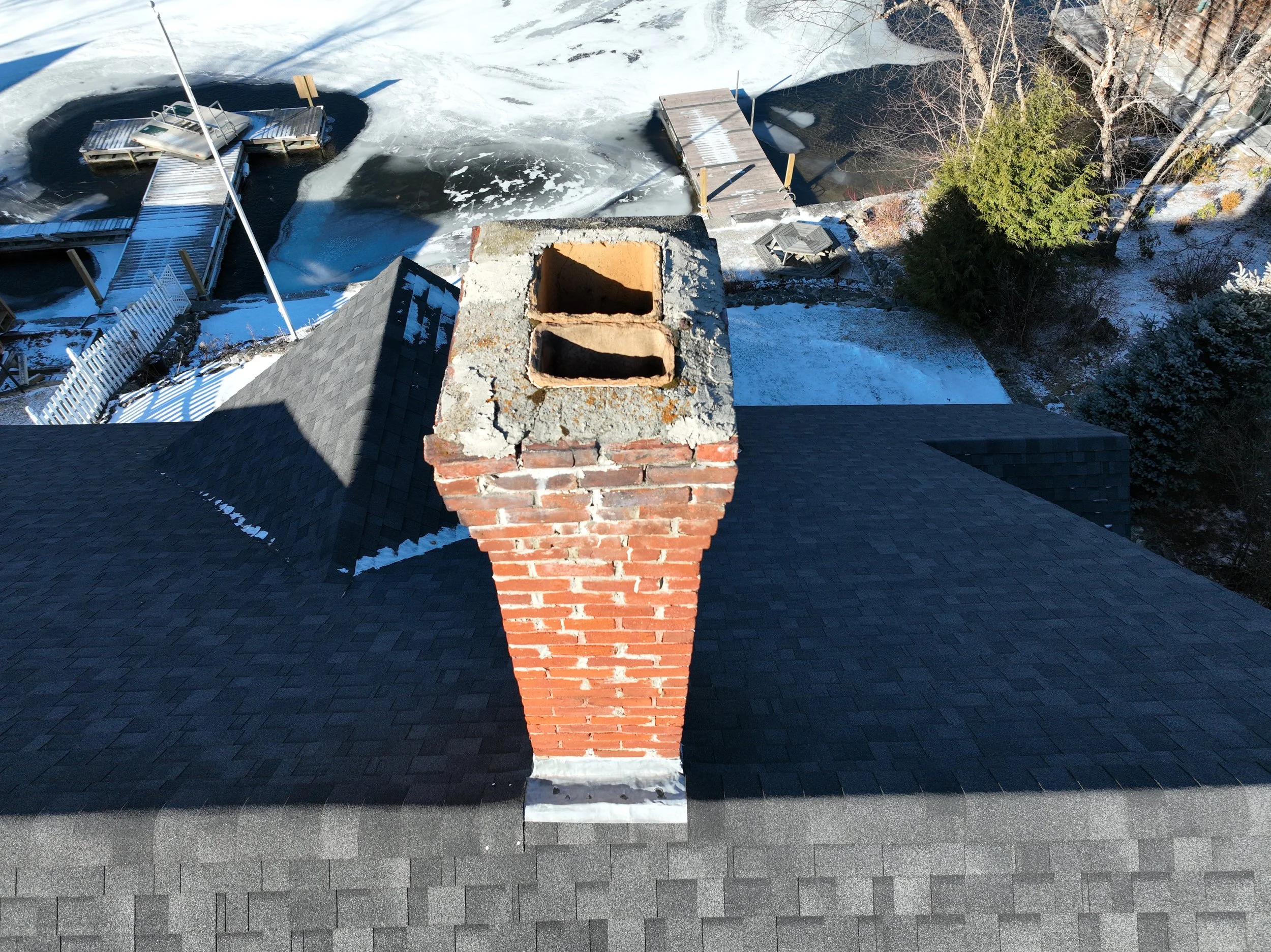 Overhead view of a house roof with a brick chimney and surrounding snow-covered landscape, including trees and a frozen pond with docks.