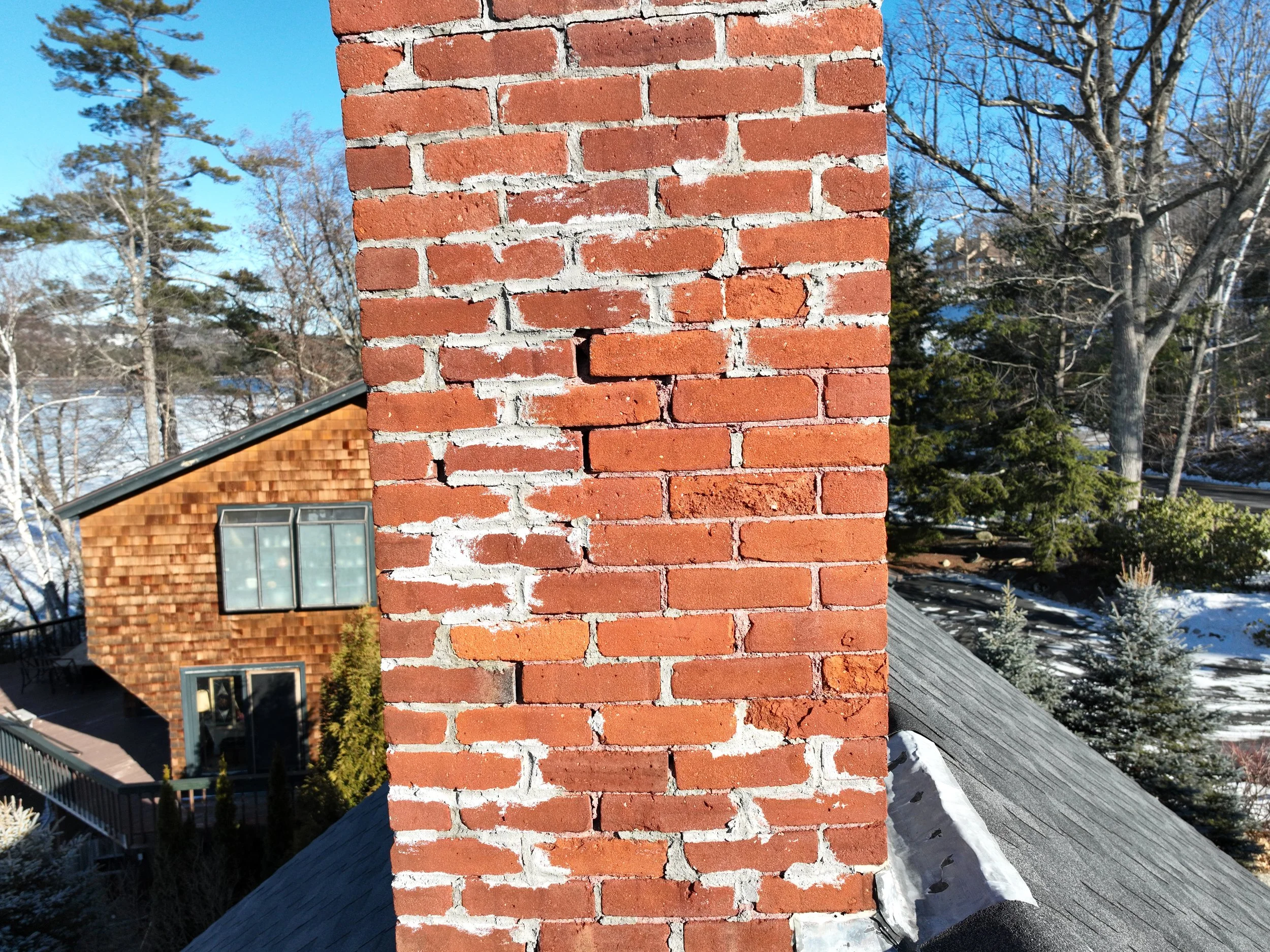 Close-up of a brick chimney on a roof with a snowy suburban yard and trees in the background.