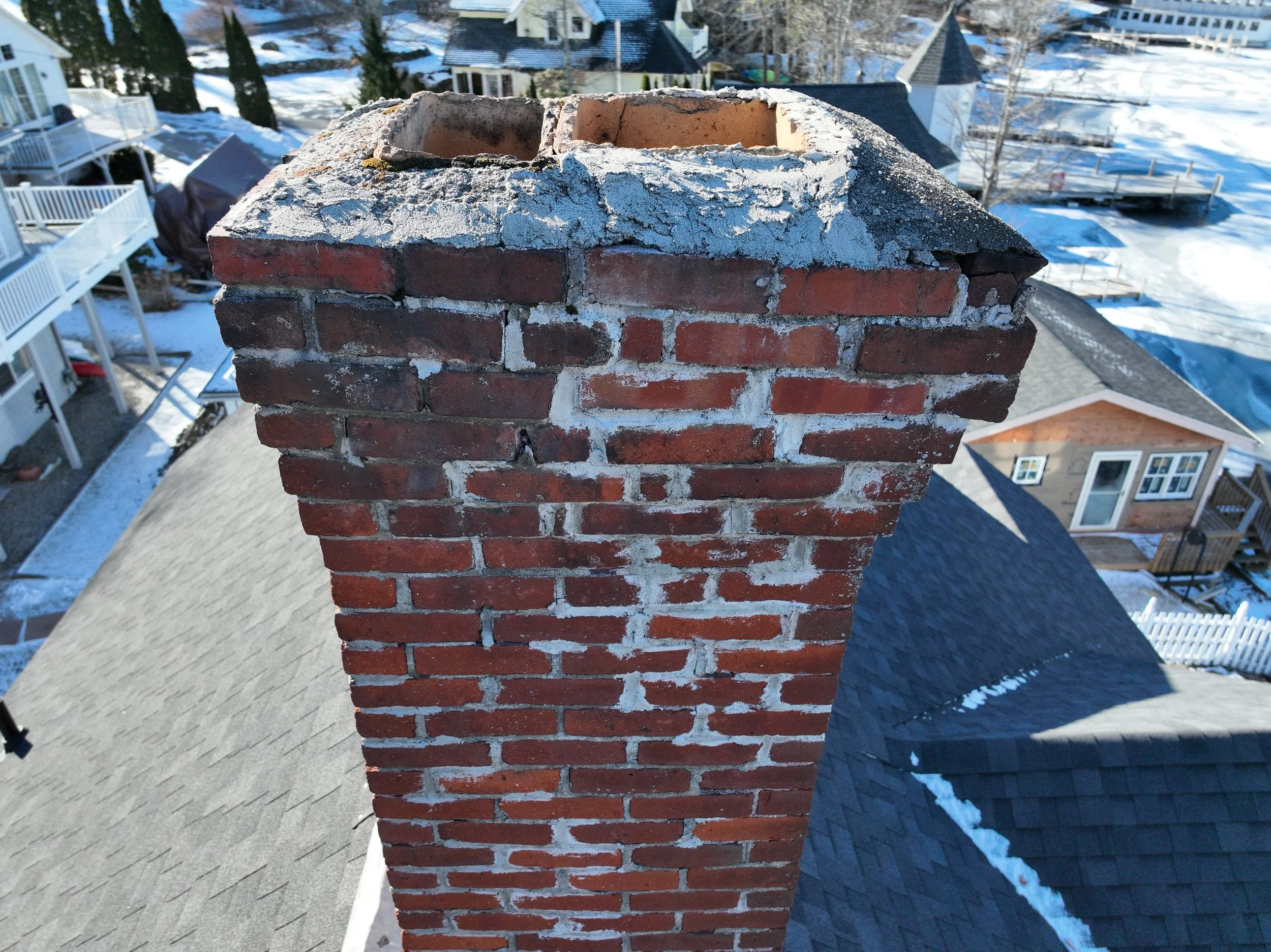Close-up view of a brick chimney on a house roof during winter, with snow visible on surrounding rooftops and ground in the background.