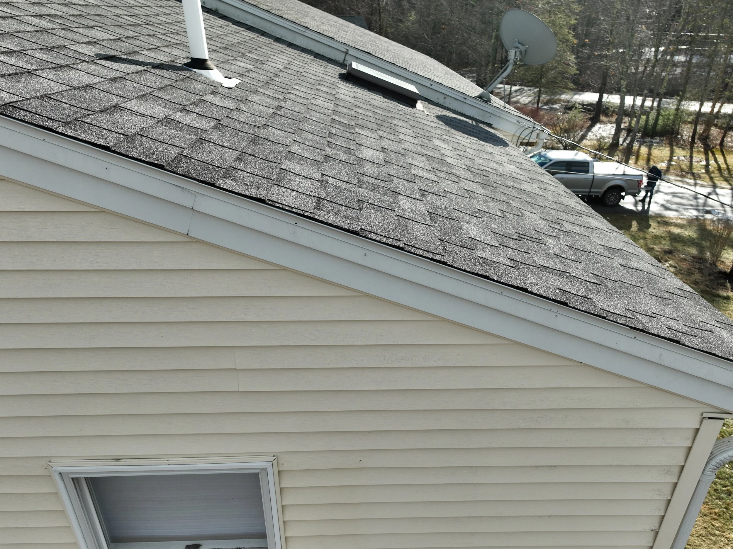 A house's beige vinyl siding wall with a window, a gray asphalt shingle roof, a white gutter, a satellite dish, and a roof vent, with a background of trees, a parked pickup truck, and a person.