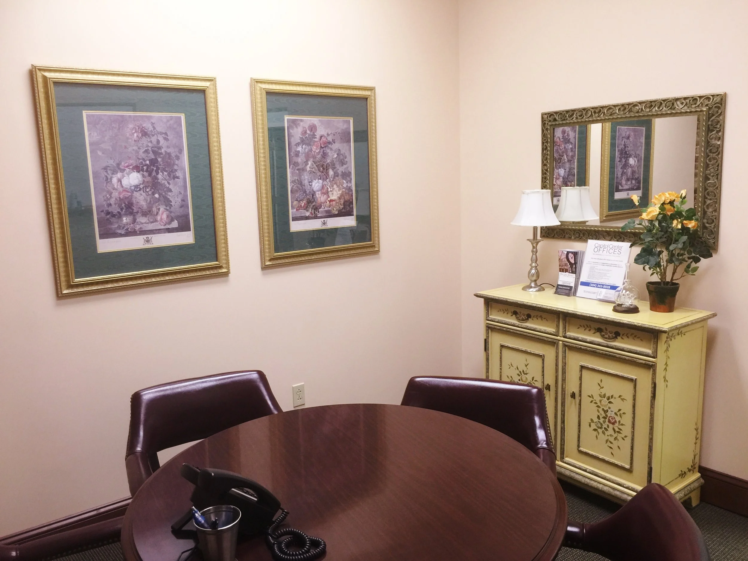 Meeting room with a round wood table, four leather chairs, a yellow painted cabinet with floral decorations, a framed mirror, and wall art of floral still life paintings.